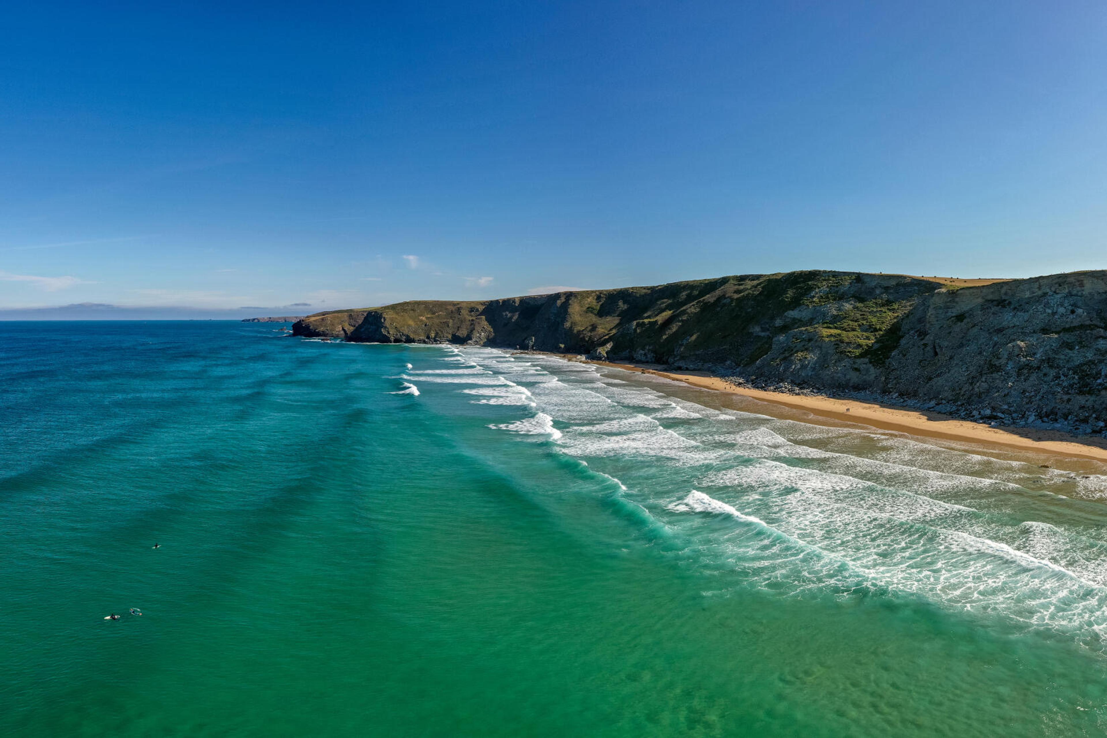 An aerial view of a curving sandy beach between headlands, with turquoise shallows giving way to deep blue sea.