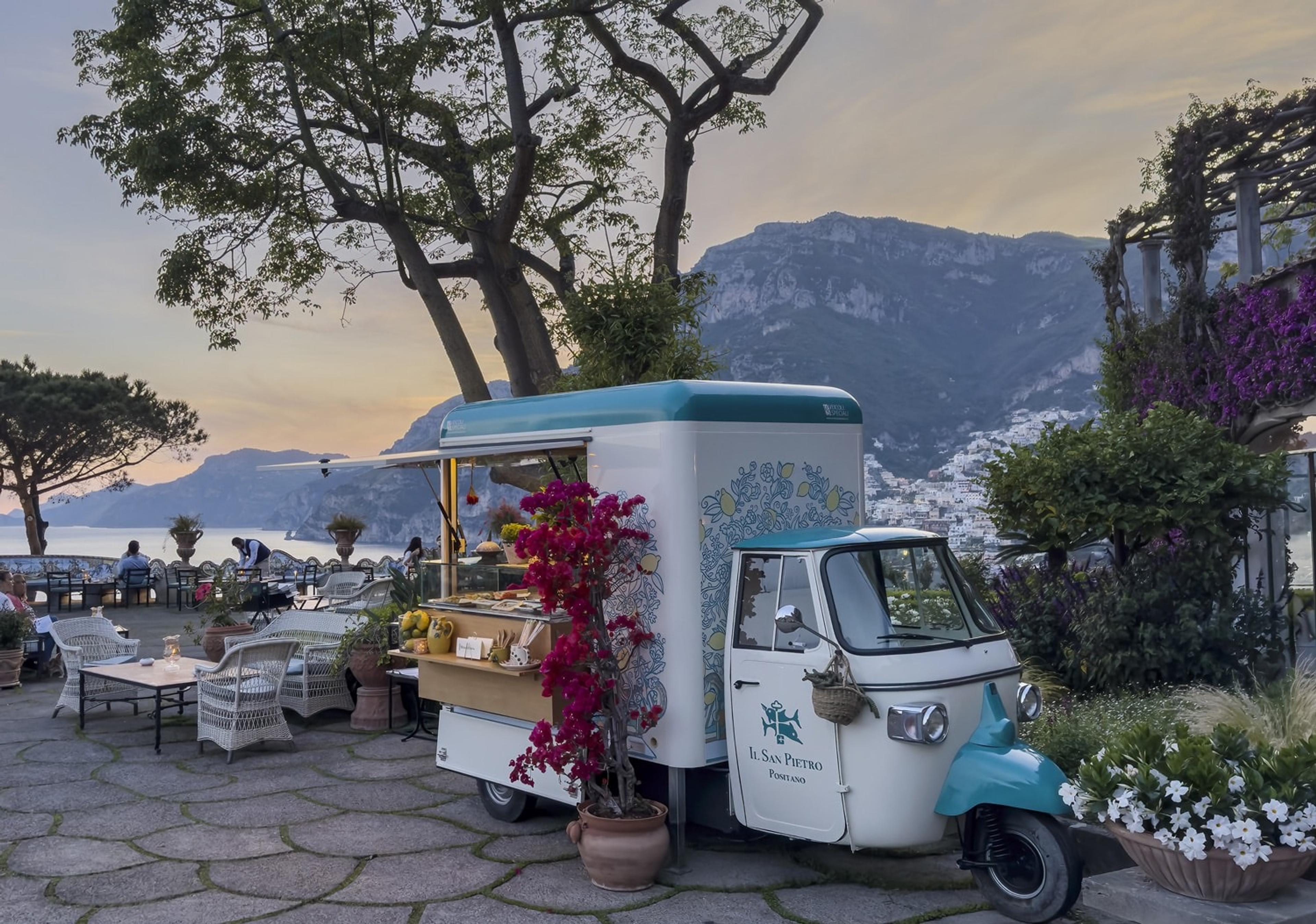 A branded three-wheeled food cart parked on a stone terrace among flowering plants, with a hillside town and sea visible at dusk.