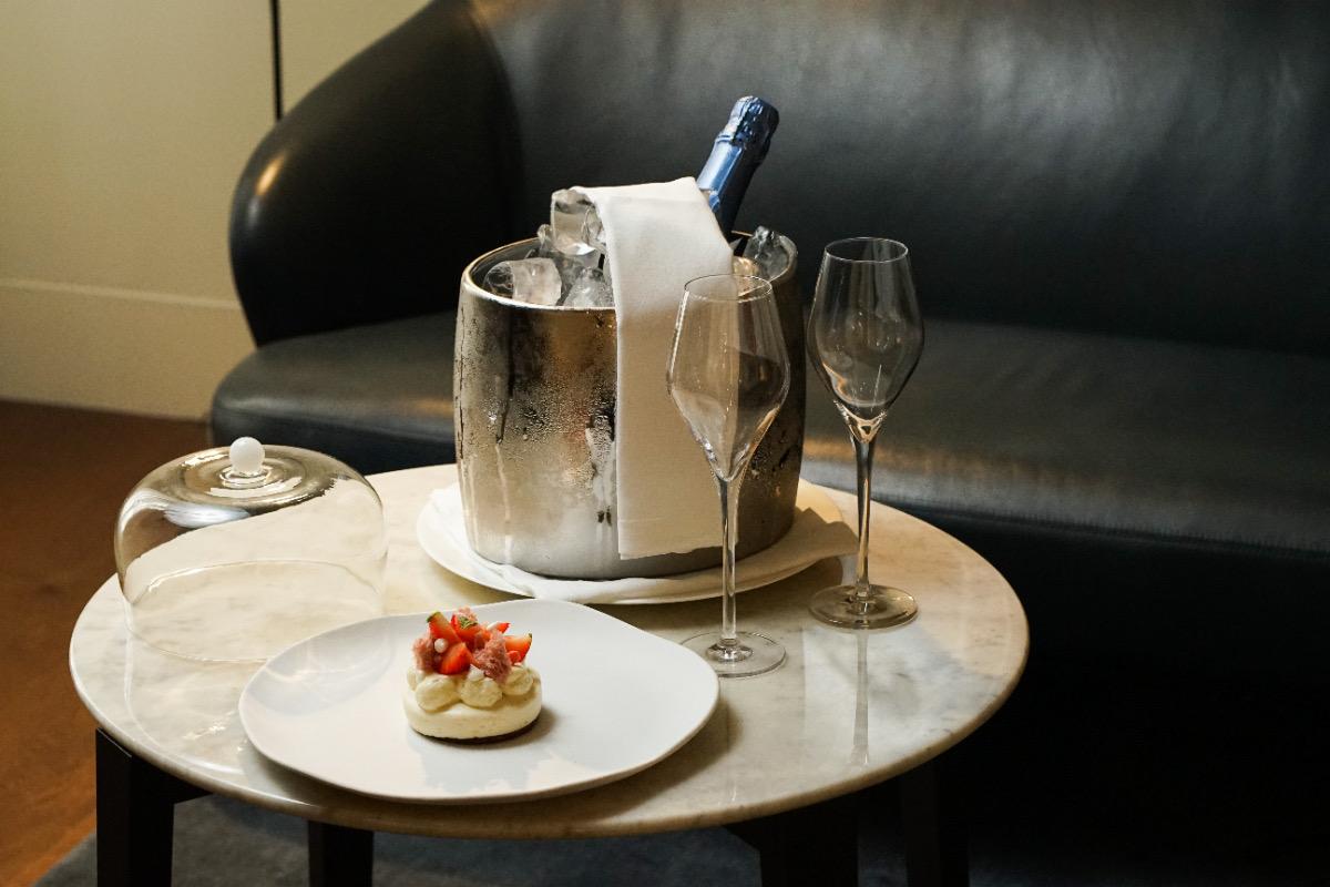 A champagne bottle chills in an ice bucket beside two flutes and a small tart on a plate, set on a marble table in front of a dark leather sofa.