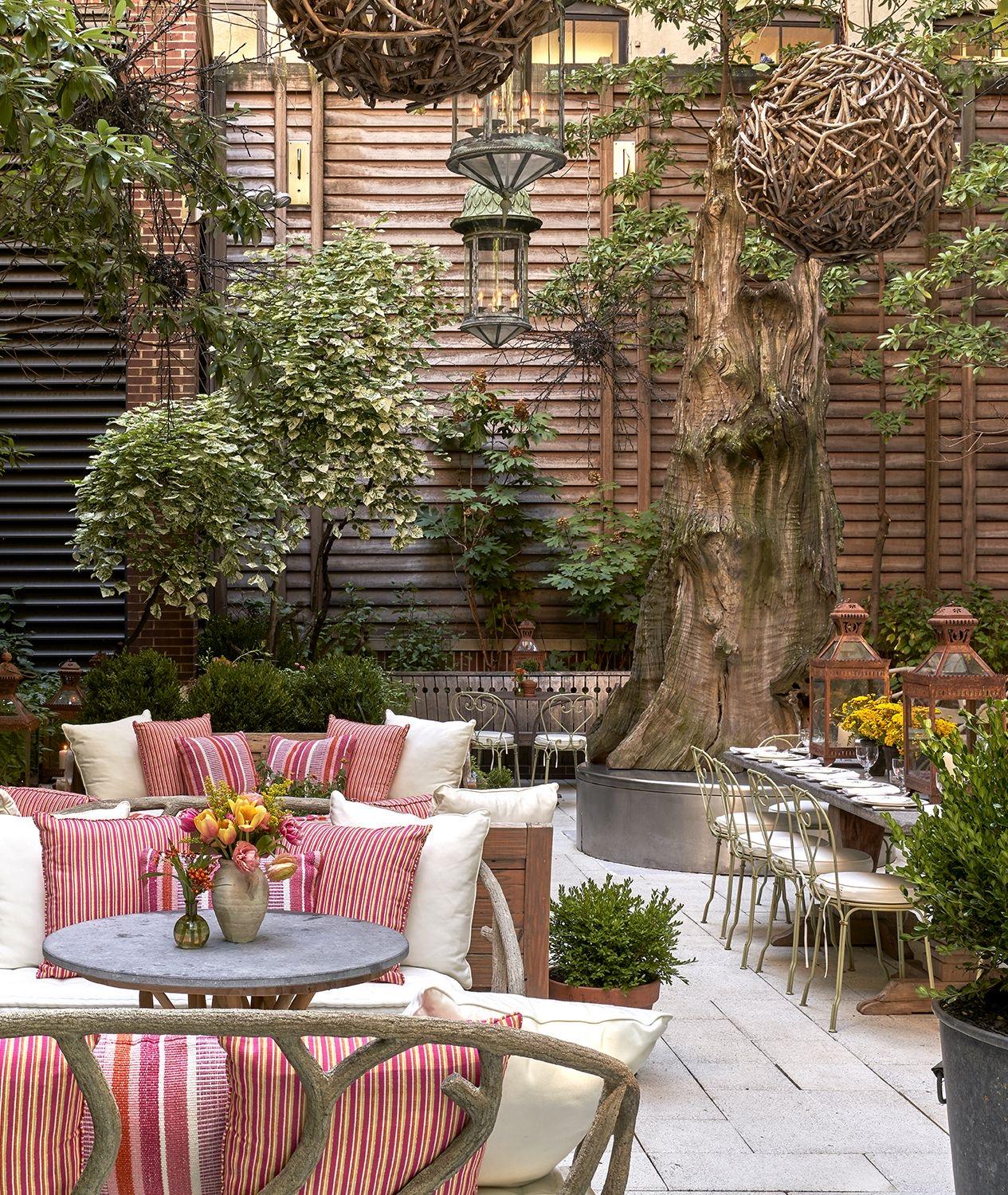 A courtyard seating area with pink and white striped cushions around a low round table holding a jug of flowers, with a long dining table set beyond it under a large bare tree trunk hung with woven lanterns.