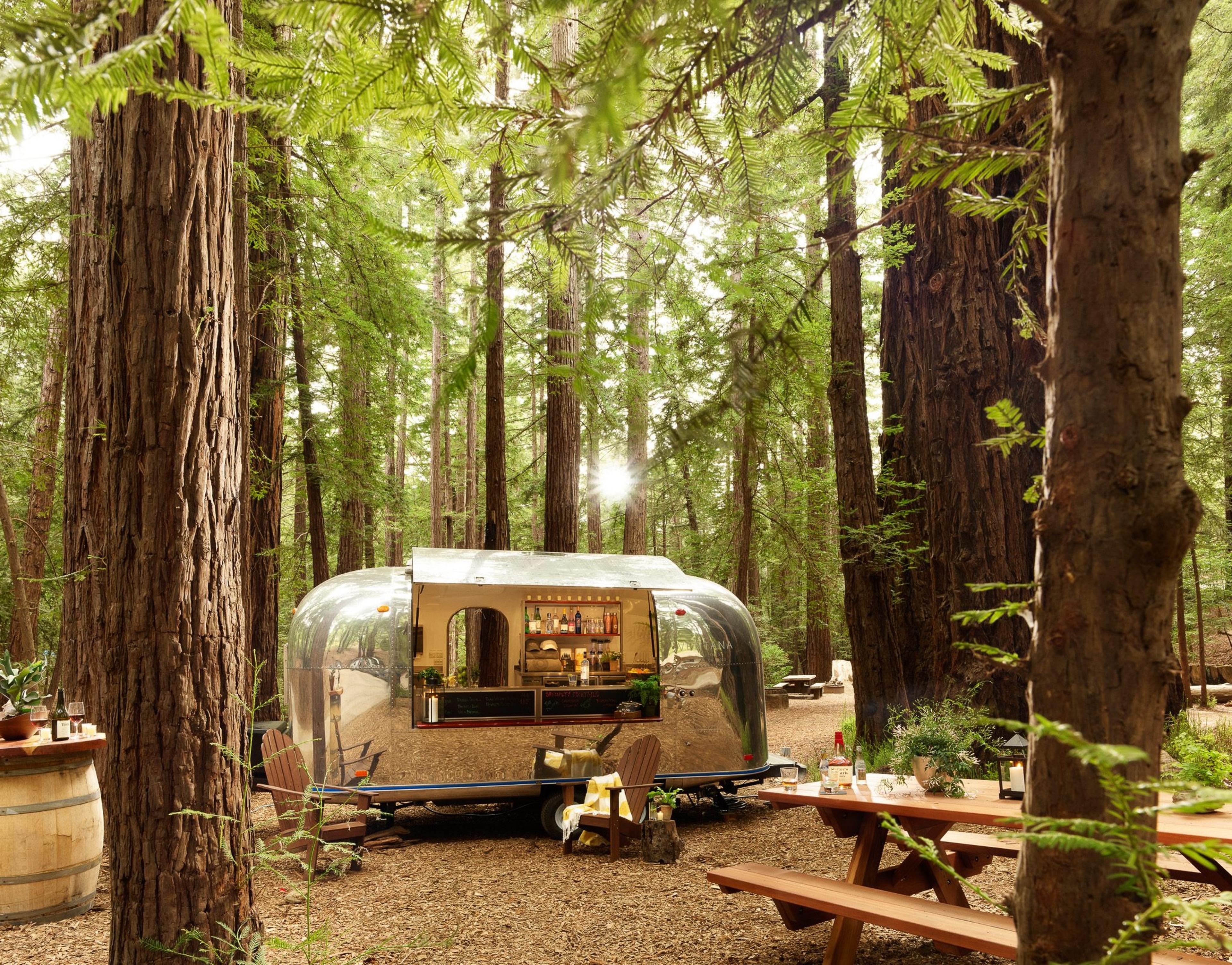 a polished silver Airstream trailer set up as a bar among redwood trees, with picnic tables, wine bottles and glasses in the foreground