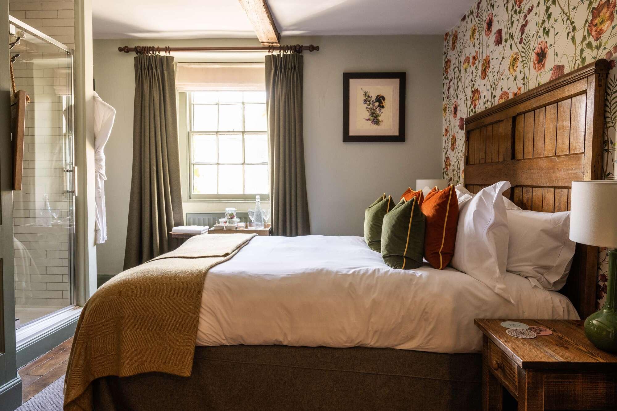 A bedroom with a dark wooden headboard, green and white floral wallpaper, bedside lamps and curtains either side of a window, with an open door showing a bathroom.