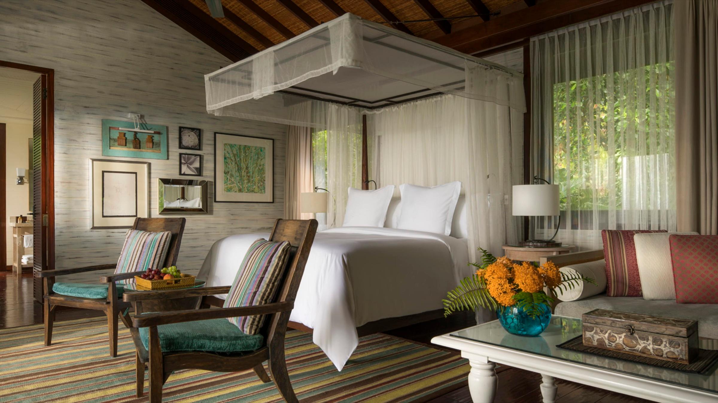 A bedroom with a sheer canopy over the bed, two striped armchairs and a vase of orange flowers on the coffee table.