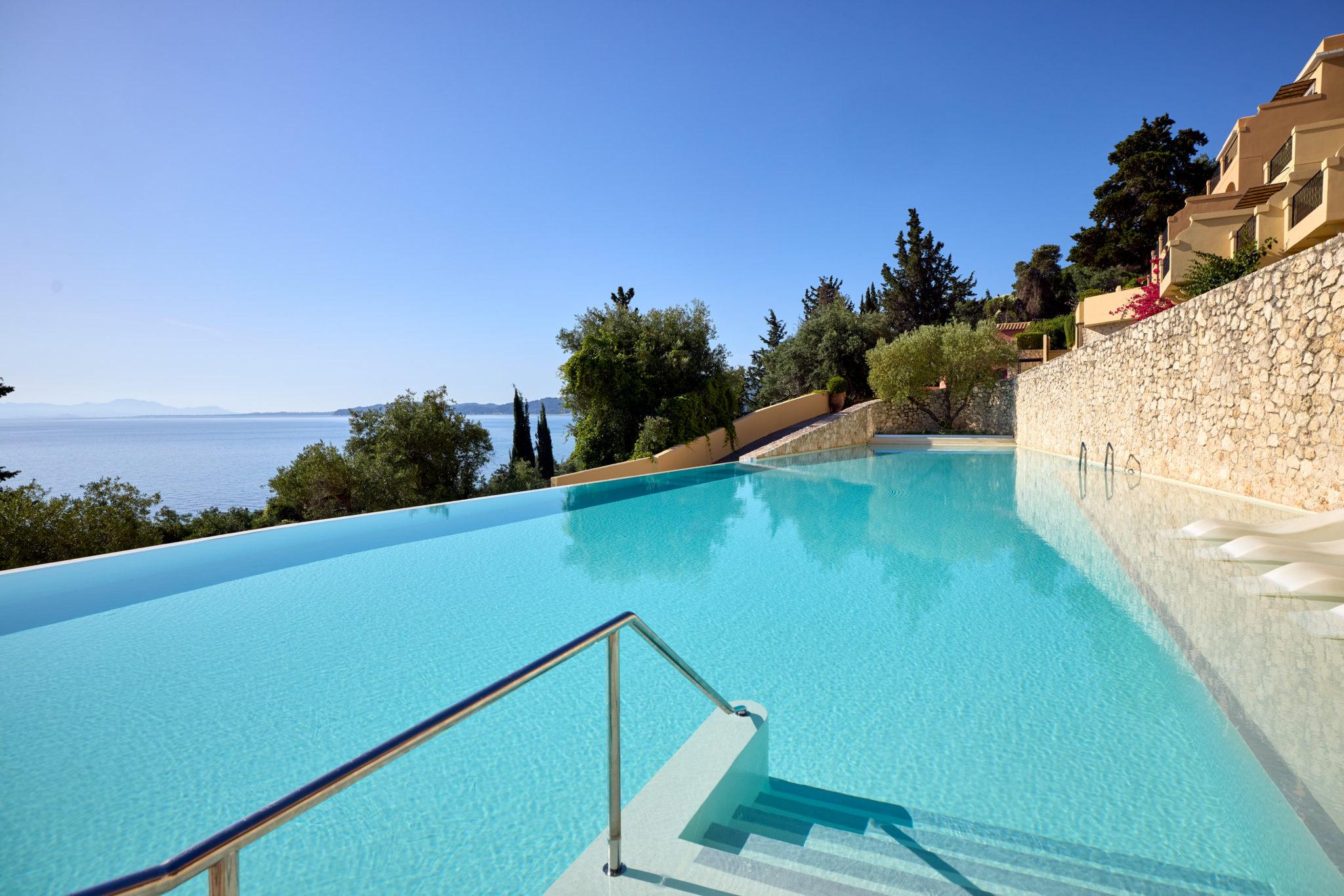 A wide infinity pool bordered by a stone wall and trees, with the sea, hills and a corner of the hotel building visible beyond.