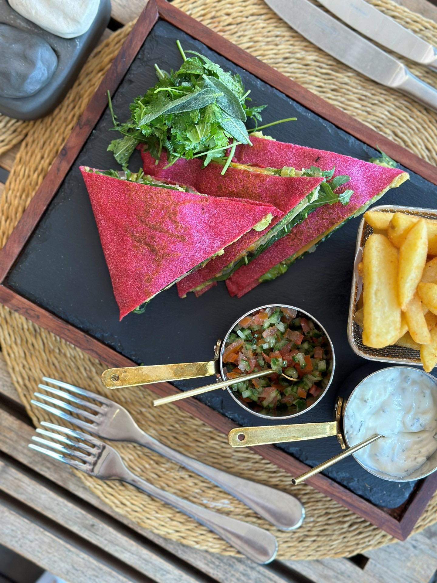 Two folded flatbread sandwiches filled with fried chicken, slaw and herbs on a dark plate, with a basket of fries alongside.