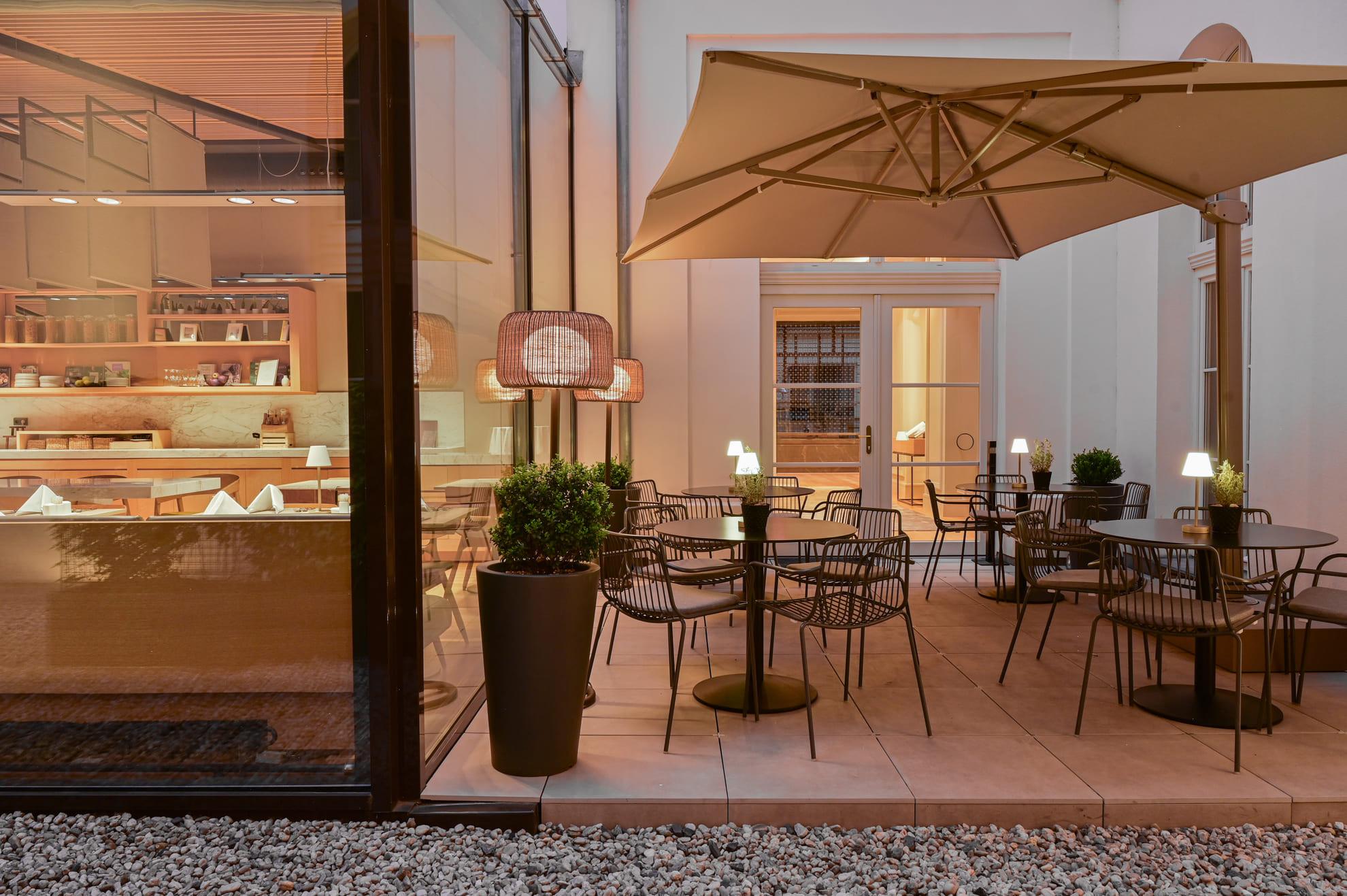 An outdoor terrace under a large cream parasol, with dark metal tables and chairs on pale tiles, and a glass-fronted room with shelves lit behind it.
