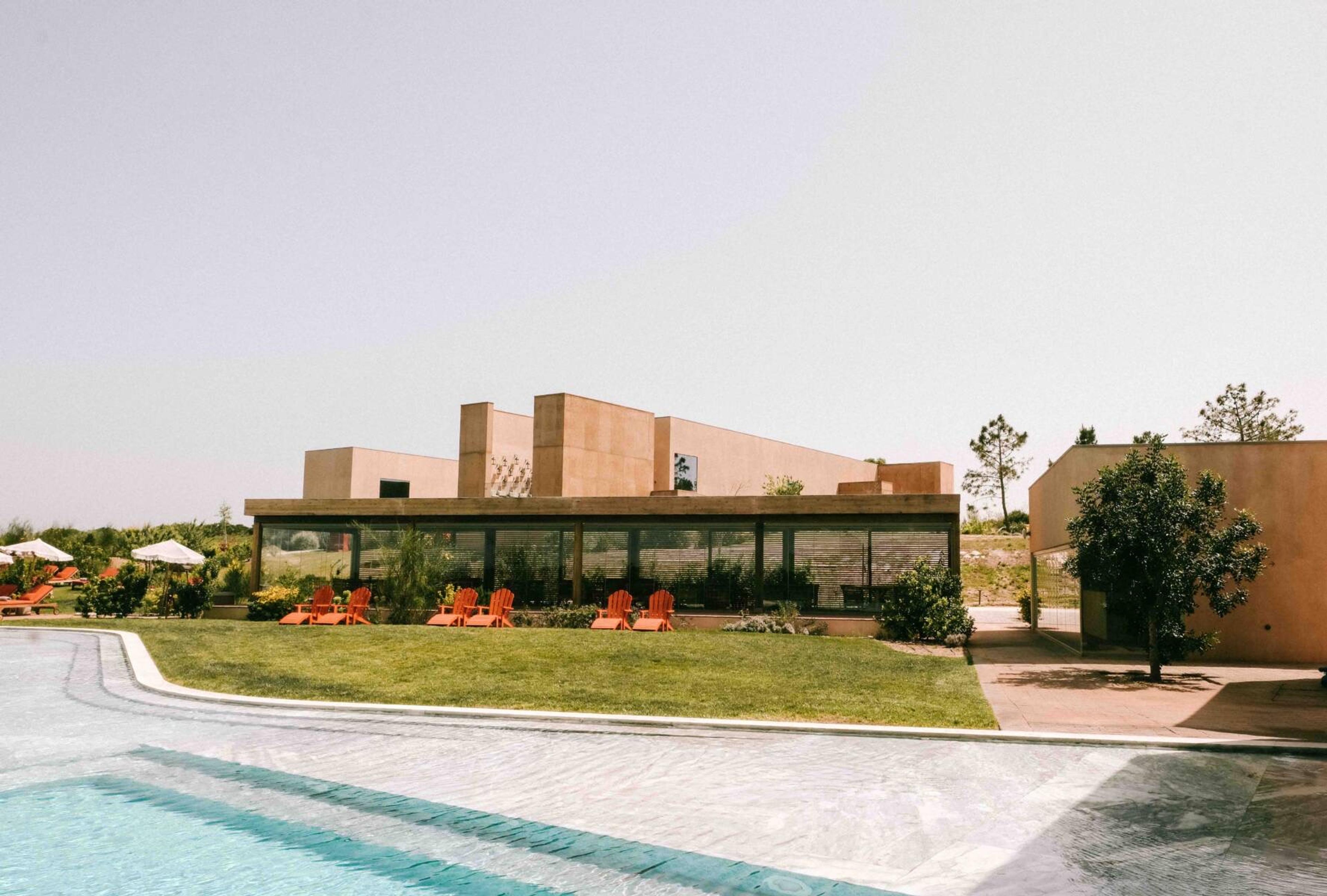 A single-storey building beside a long infinity pool, with orange sunloungers on the lawn and pine trees behind.