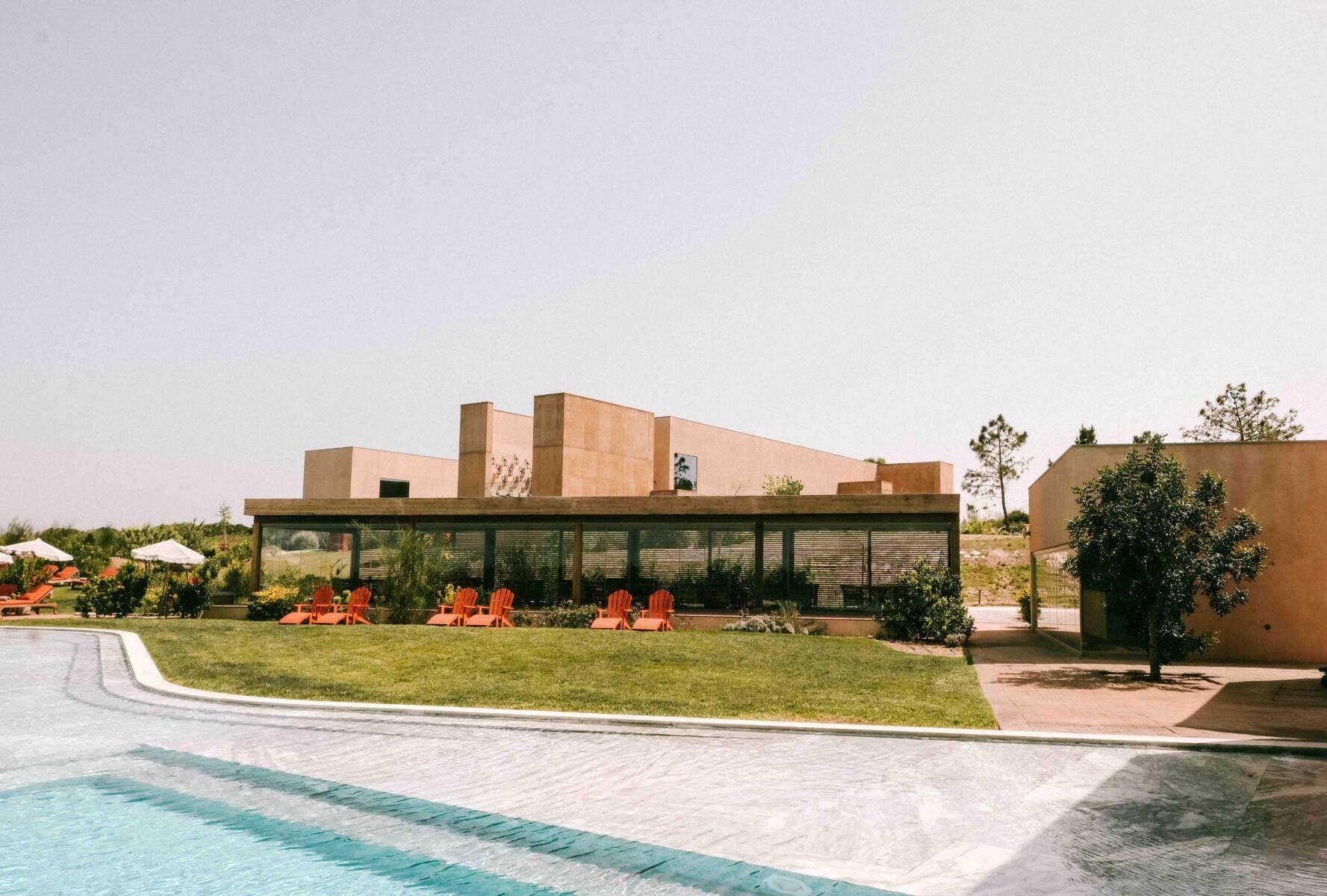 A single-storey building beside a long infinity pool, with orange sunloungers on the lawn and pine trees behind.