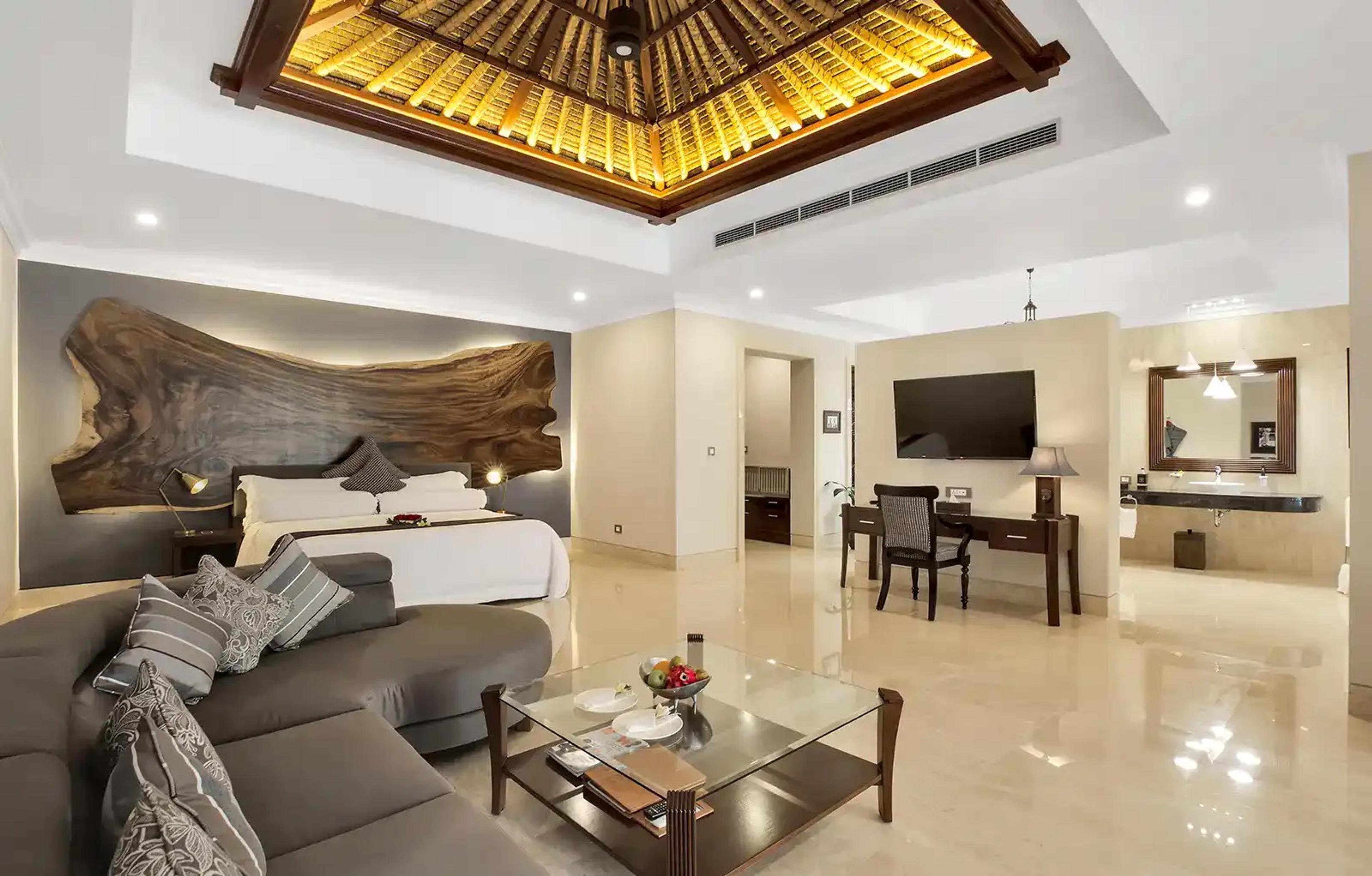 A bedroom with a raw wood slab headboard and a grey sofa opens onto a desk, television and an ensuite bathroom.