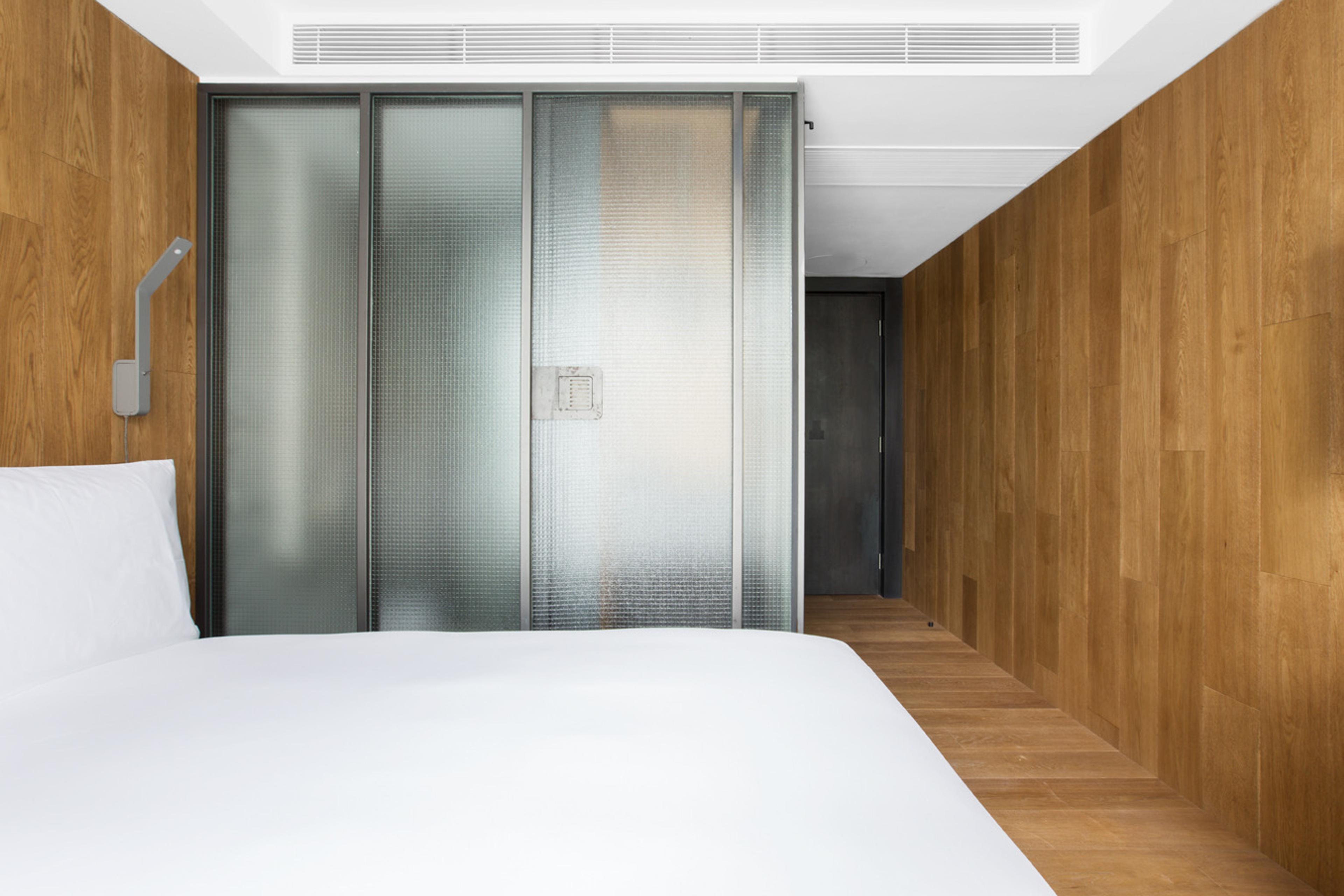 A bed in the foreground beside a frosted glass partition, looking down a wood panelled corridor to a dark door.