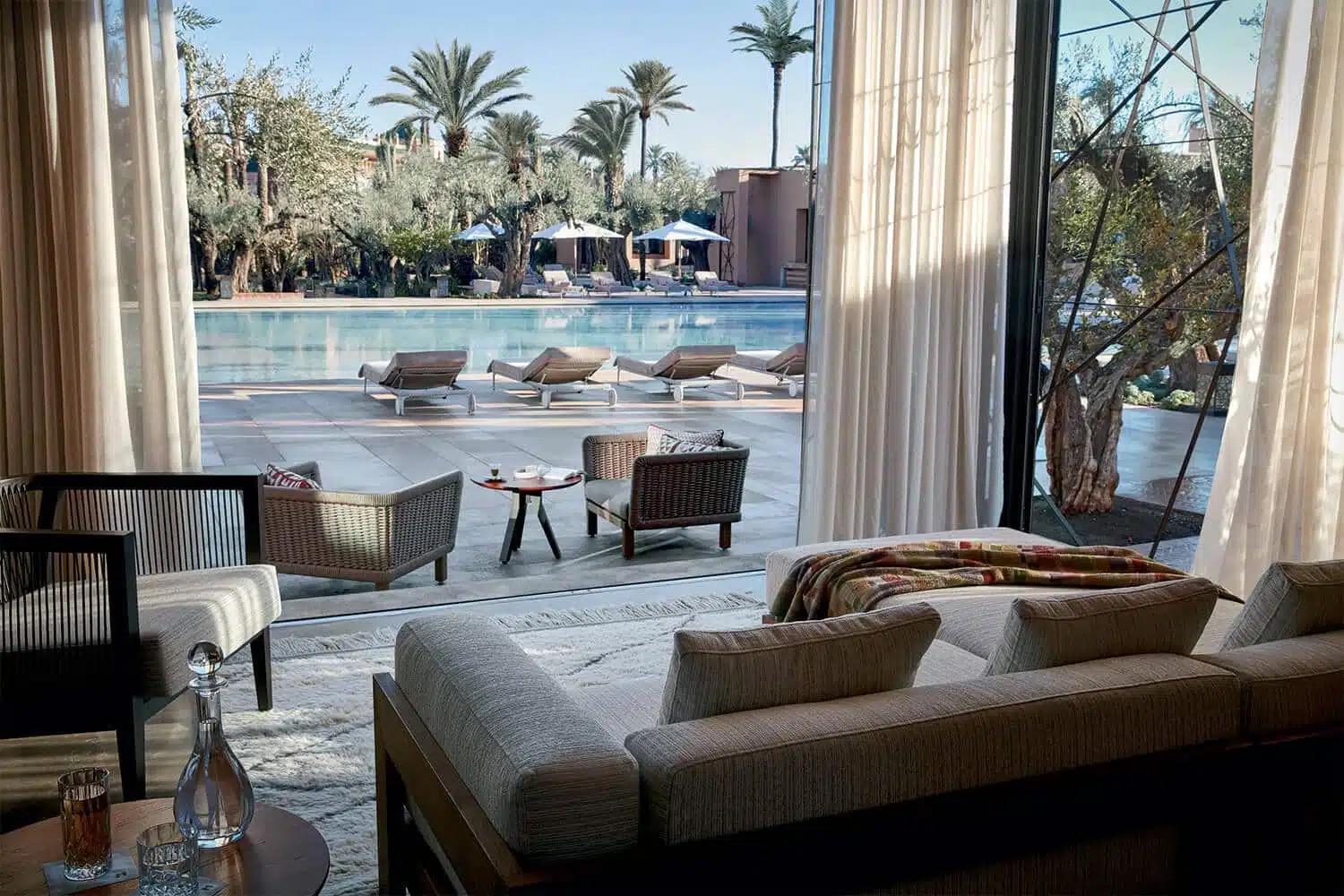A lounge with sofas and curtains opens onto a pool terrace with loungers, umbrellas and palm trees.