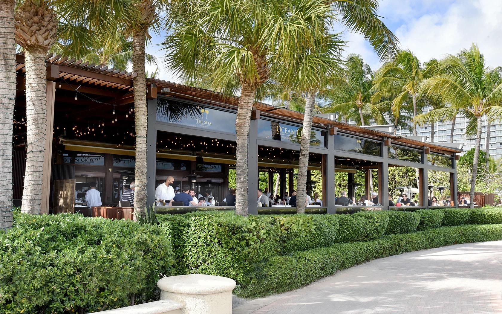 The covered, palm-shaded exterior of the Ocean Grill restaurant by day, with diners seated under string lights and a high-rise beyond.
