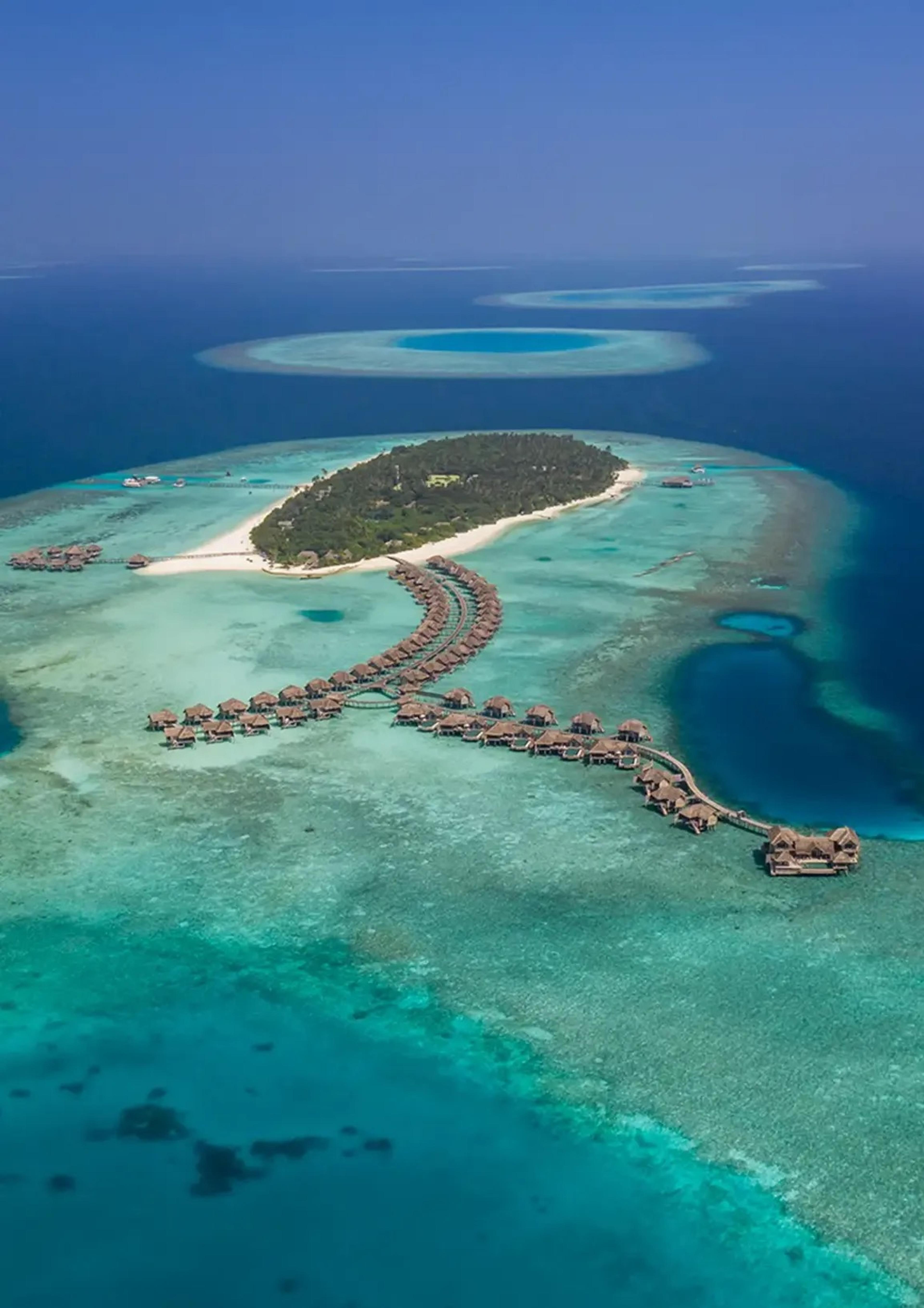 an aerial view of a resort island with overwater villas on wooden jetties, surrounded by a turquoise lagoon and coral reef, with two atoll rings visible on the horizon