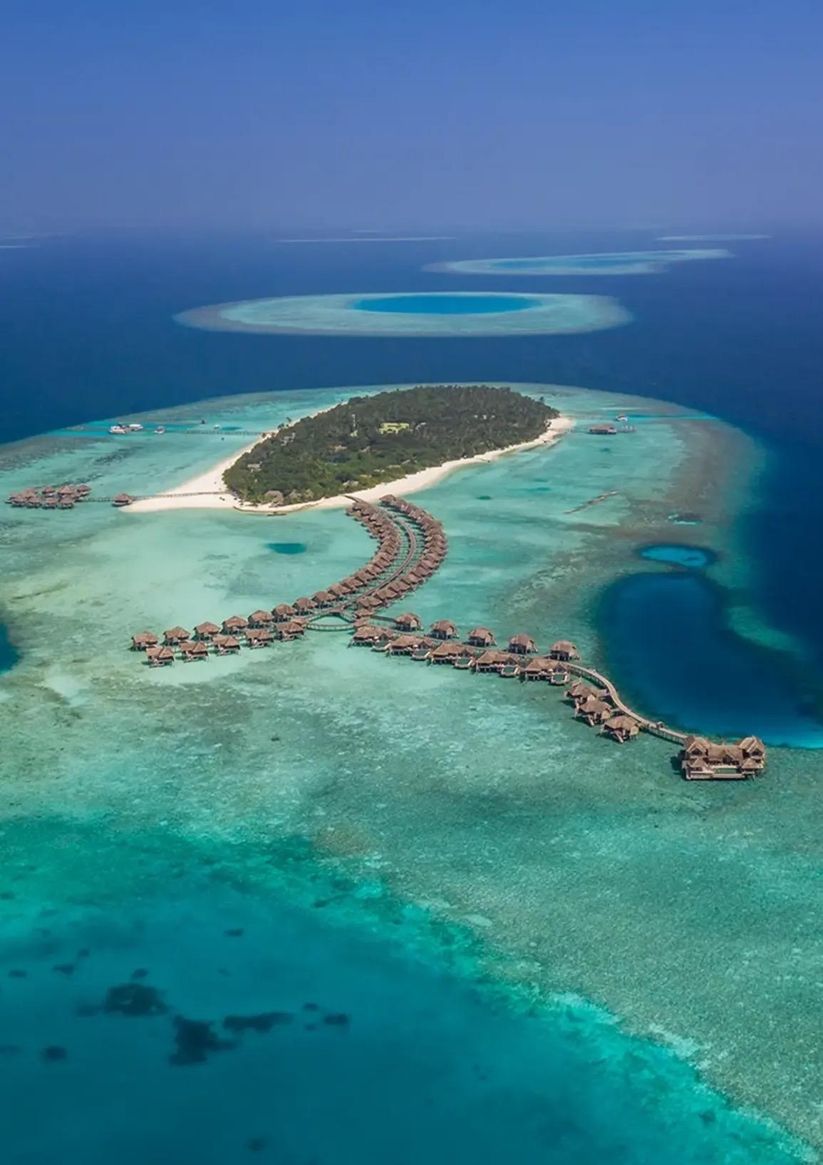 an aerial view of a resort island with overwater villas on wooden jetties, surrounded by a turquoise lagoon and coral reef, with two atoll rings visible on the horizon