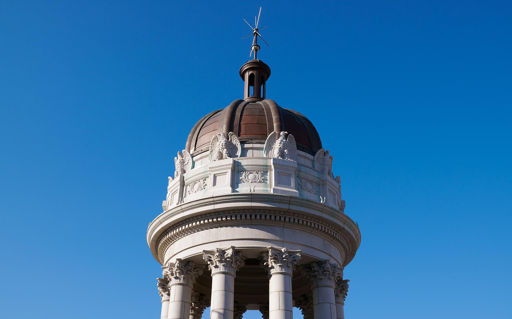 A close-up of a stone tower's domed cupola with carved eagles and a weathervane, against a clear blue sky.