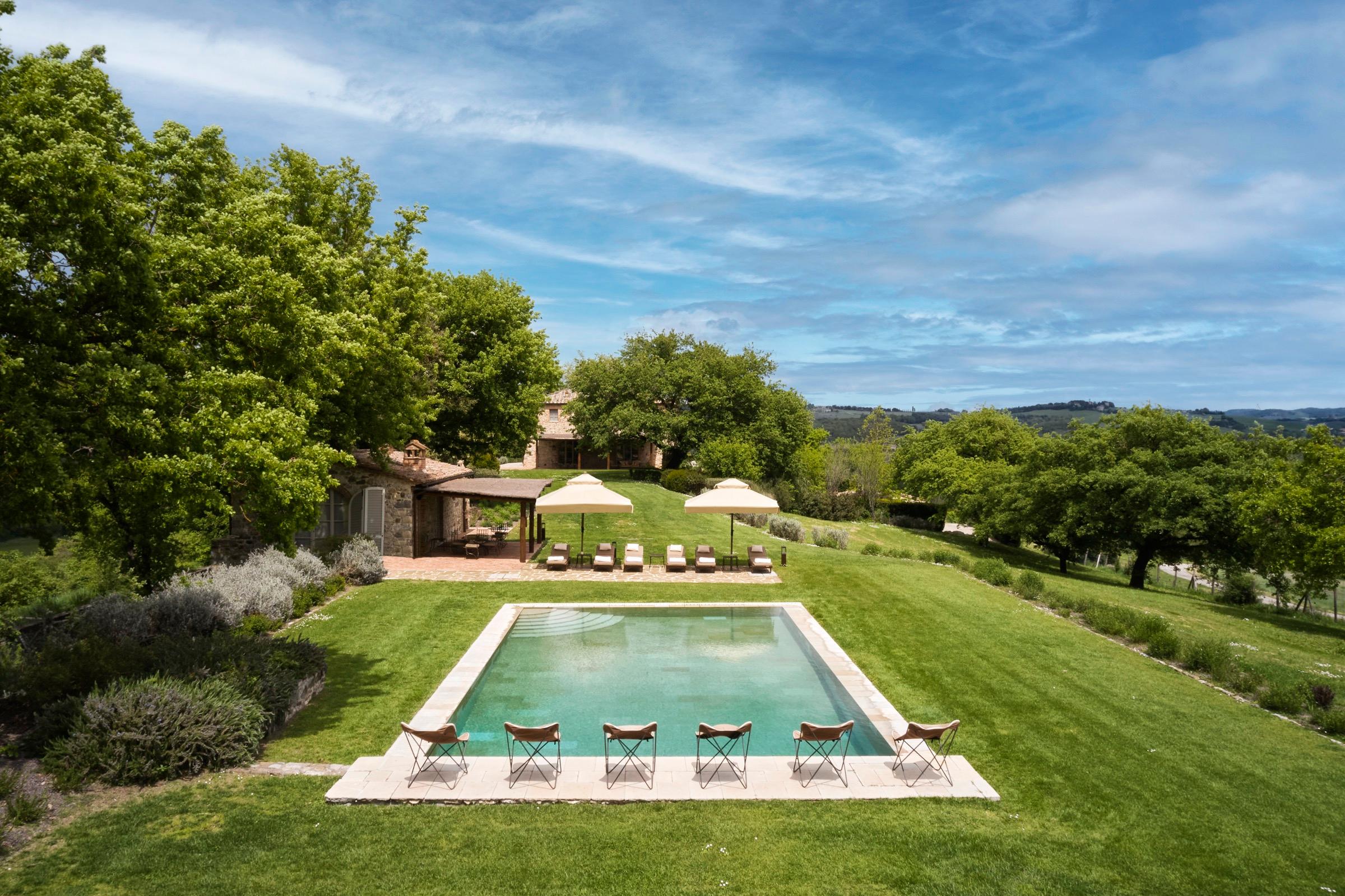 A rectangular swimming pool on a lawn beside a stone cottage, with umbrellas and loungers, trees and hills around it.
