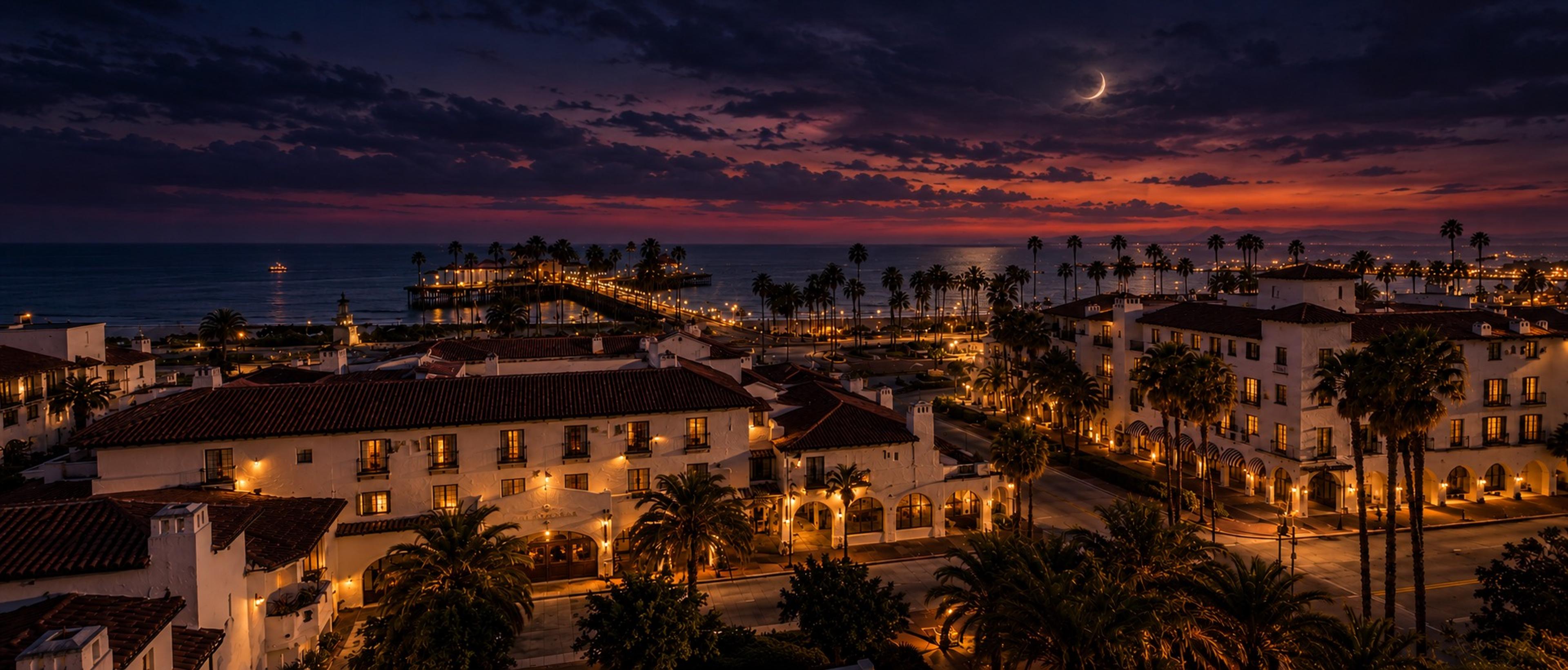 A night-time aerial view of a lit hotel complex beside a pier and the ocean, under a dark sky with a crescent moon.