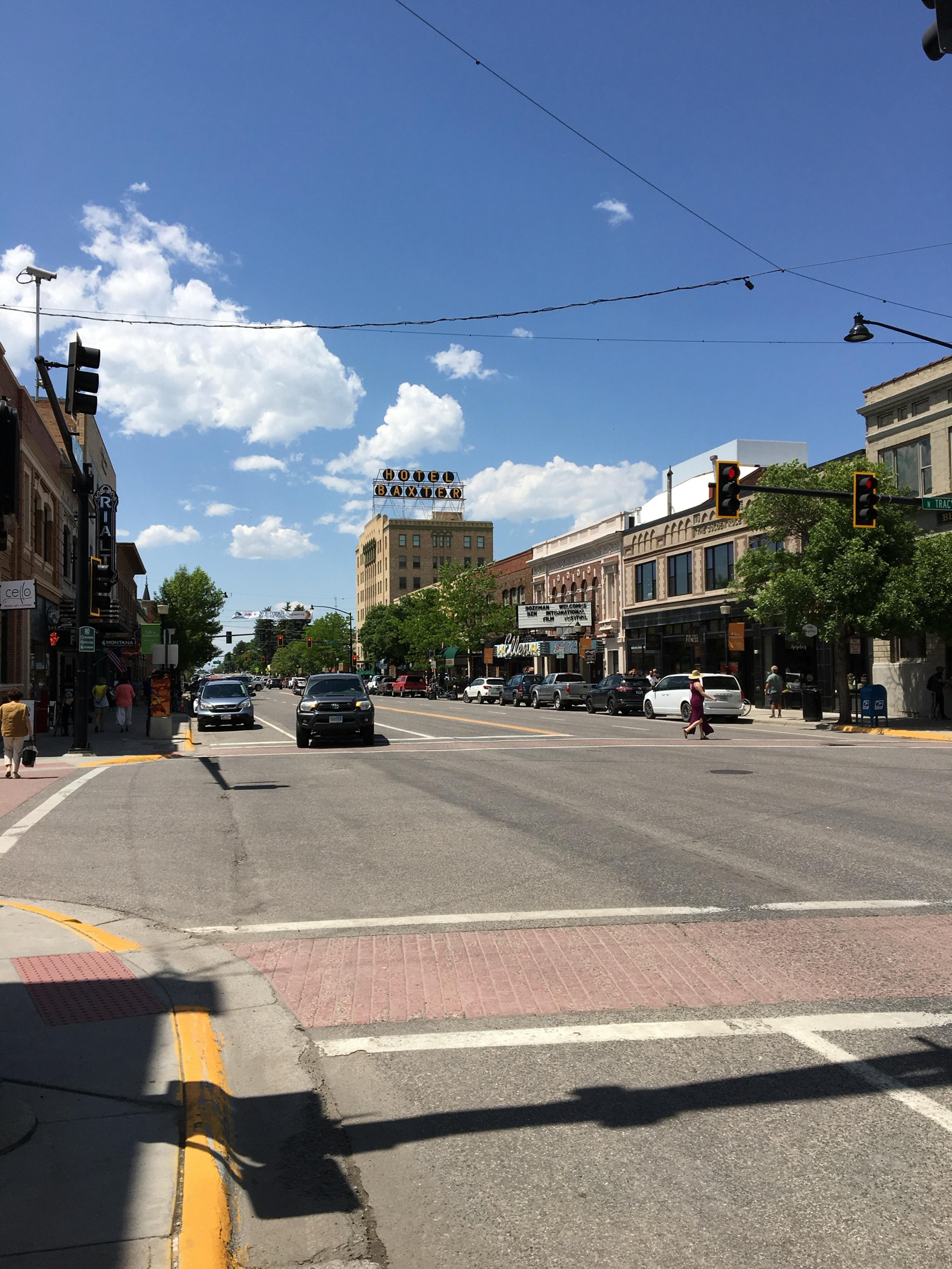 Bozeman's Main Street on a sunny afternoon, brick shopfronts on both sides and a tall hotel sign rising at the far end