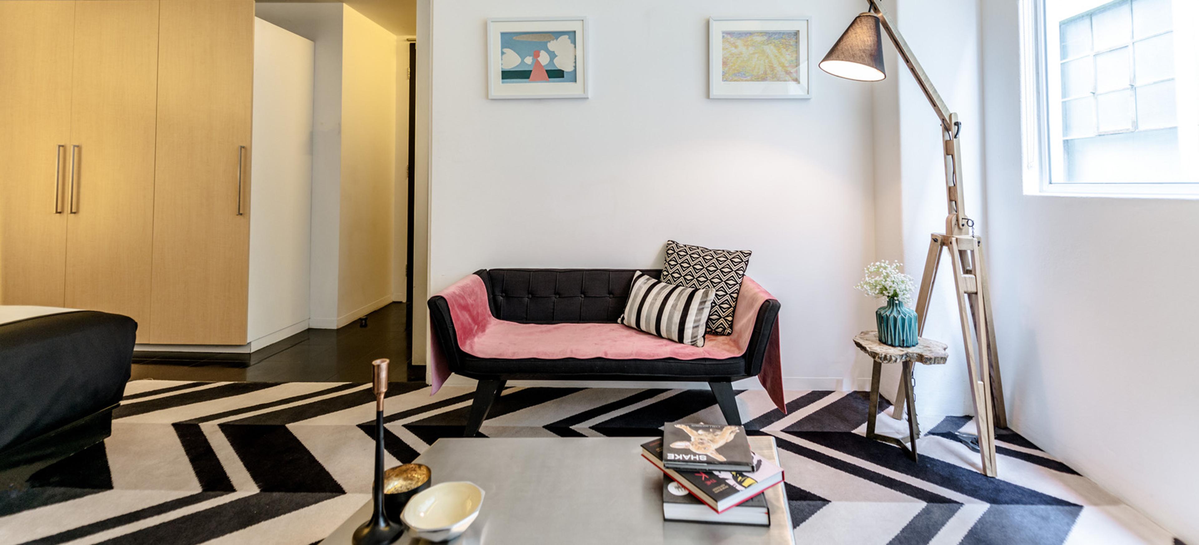 A lounge area with a black tufted daybed topped with a pink throw and patterned cushions, two framed prints on the wall, and a tripod floor lamp beside a window.