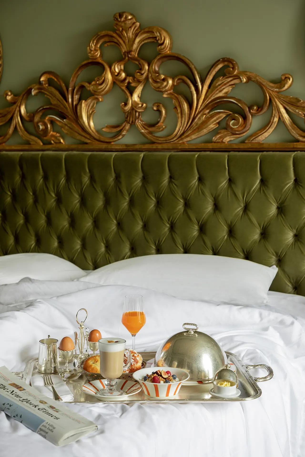 A silver breakfast tray with eggs, orange juice, coffee and a croissant sits on an unmade bed with a green tufted headboard and a folded newspaper beside it.