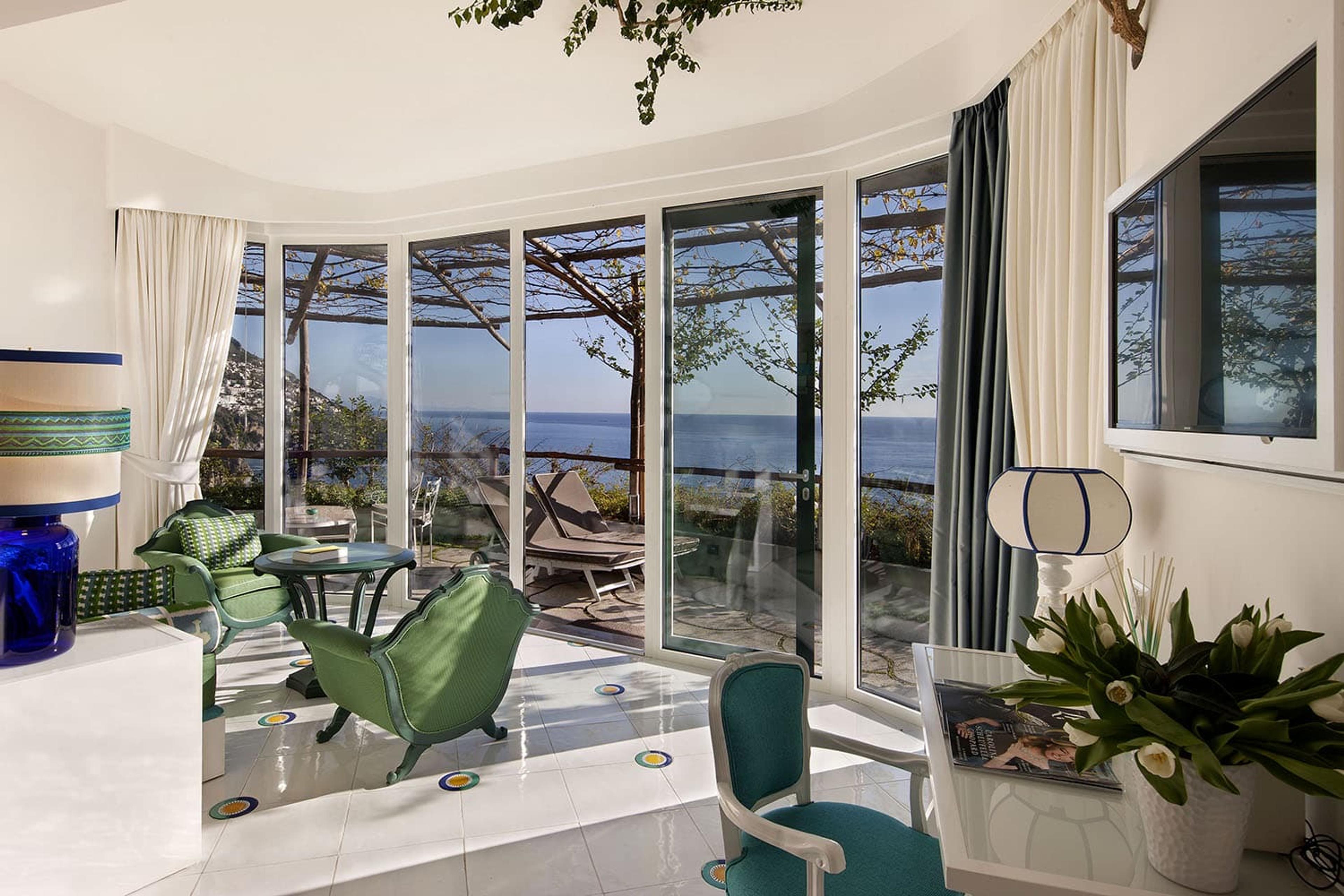 A glass-walled sitting room with green armchairs, opening onto a vine-covered terrace with sun loungers and a view of the sea.
