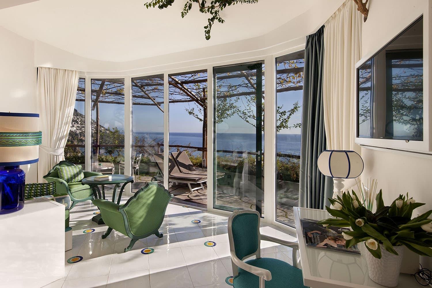 A glass-walled sitting room with green armchairs, opening onto a vine-covered terrace with sun loungers and a view of the sea.