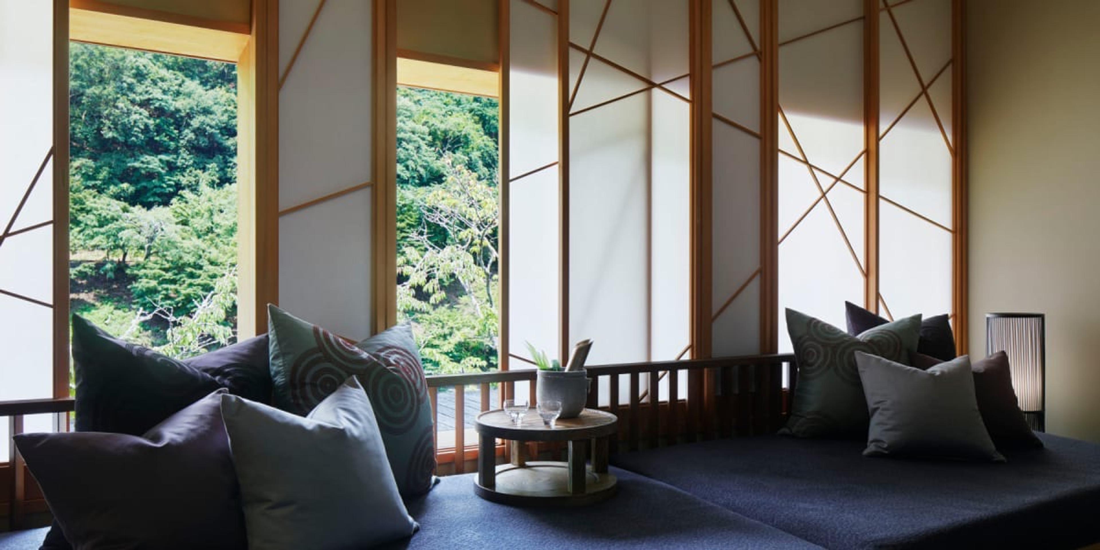 A guest room with a low sofa and cushions beside tall windows in latticed screens, forest outside