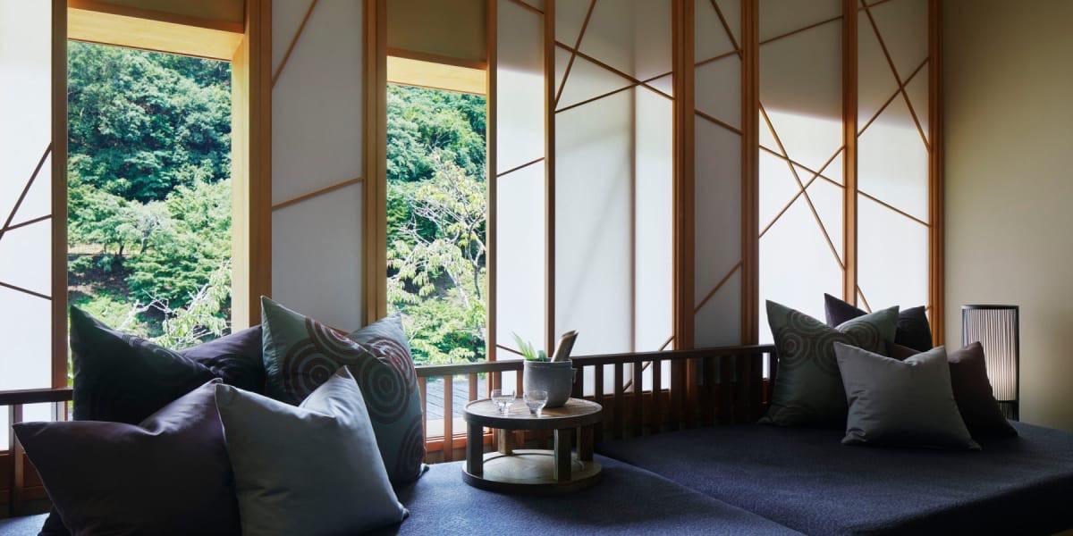 A guest room with a low sofa and cushions beside tall windows in latticed screens, forest outside