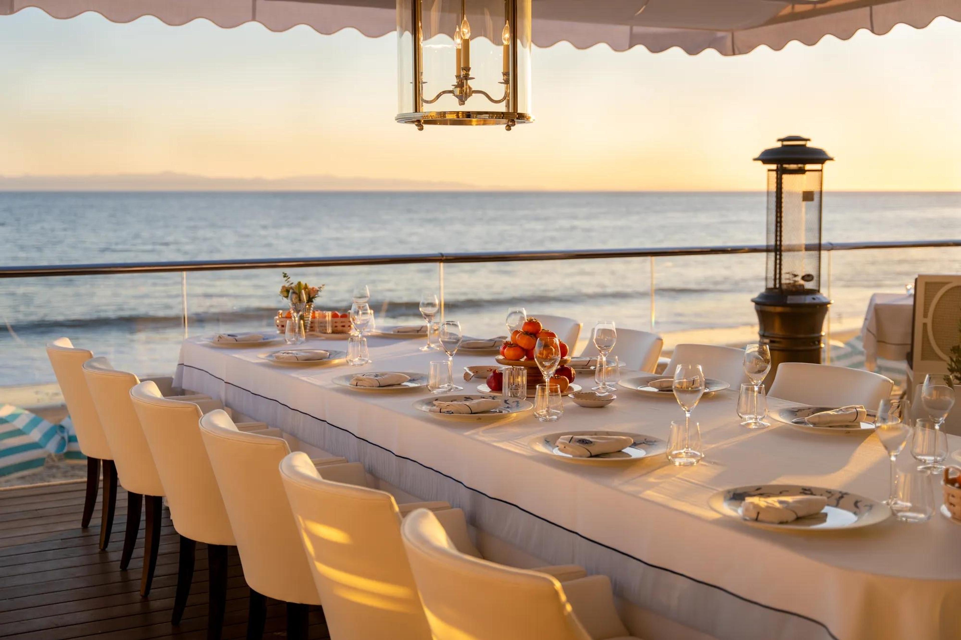 A long table set for a group dinner on a terrace, with fruit and glassware laid out and the sun setting over the ocean.