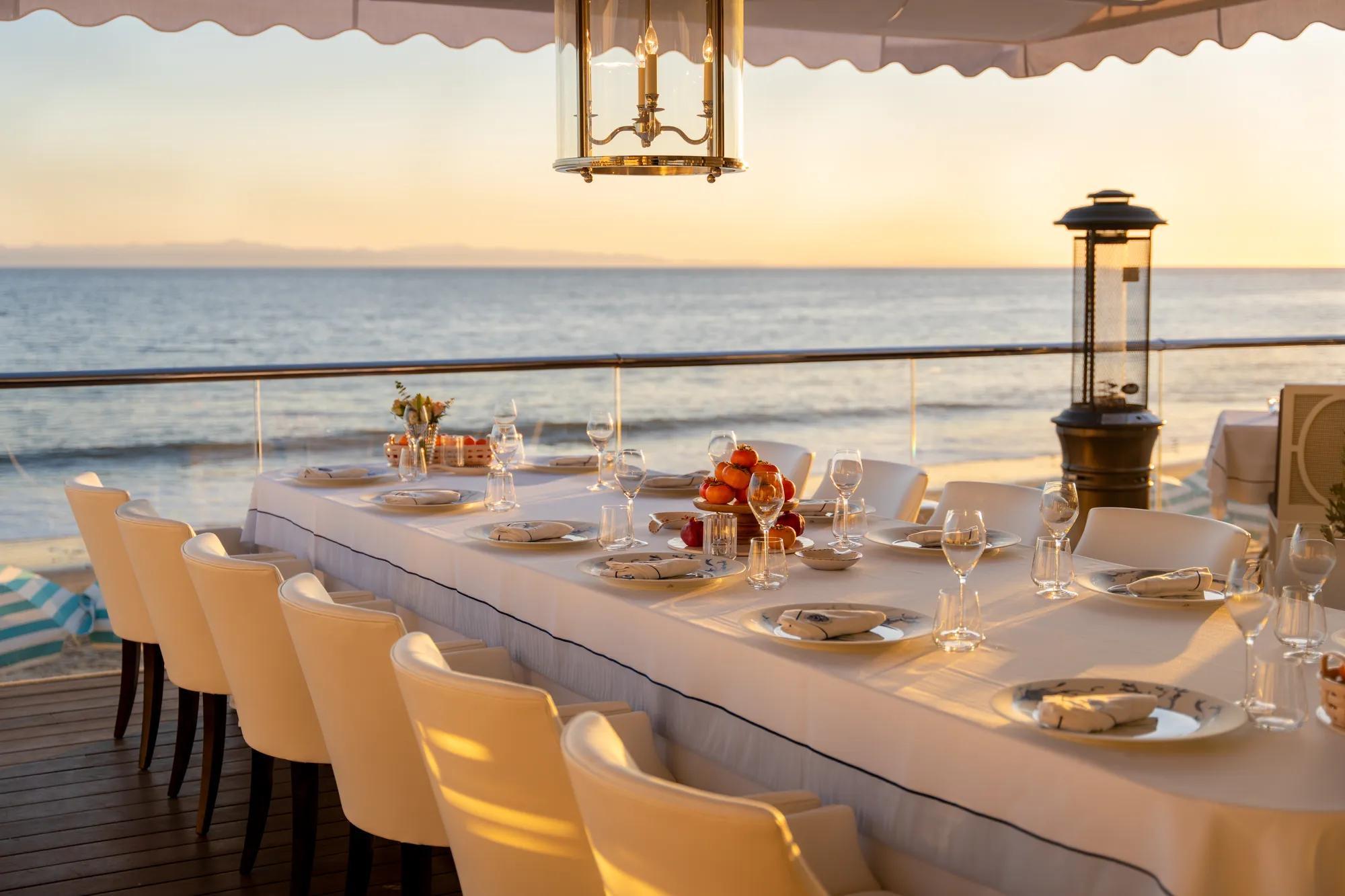 A long table set for a group dinner on a terrace, with fruit and glassware laid out and the sun setting over the ocean.