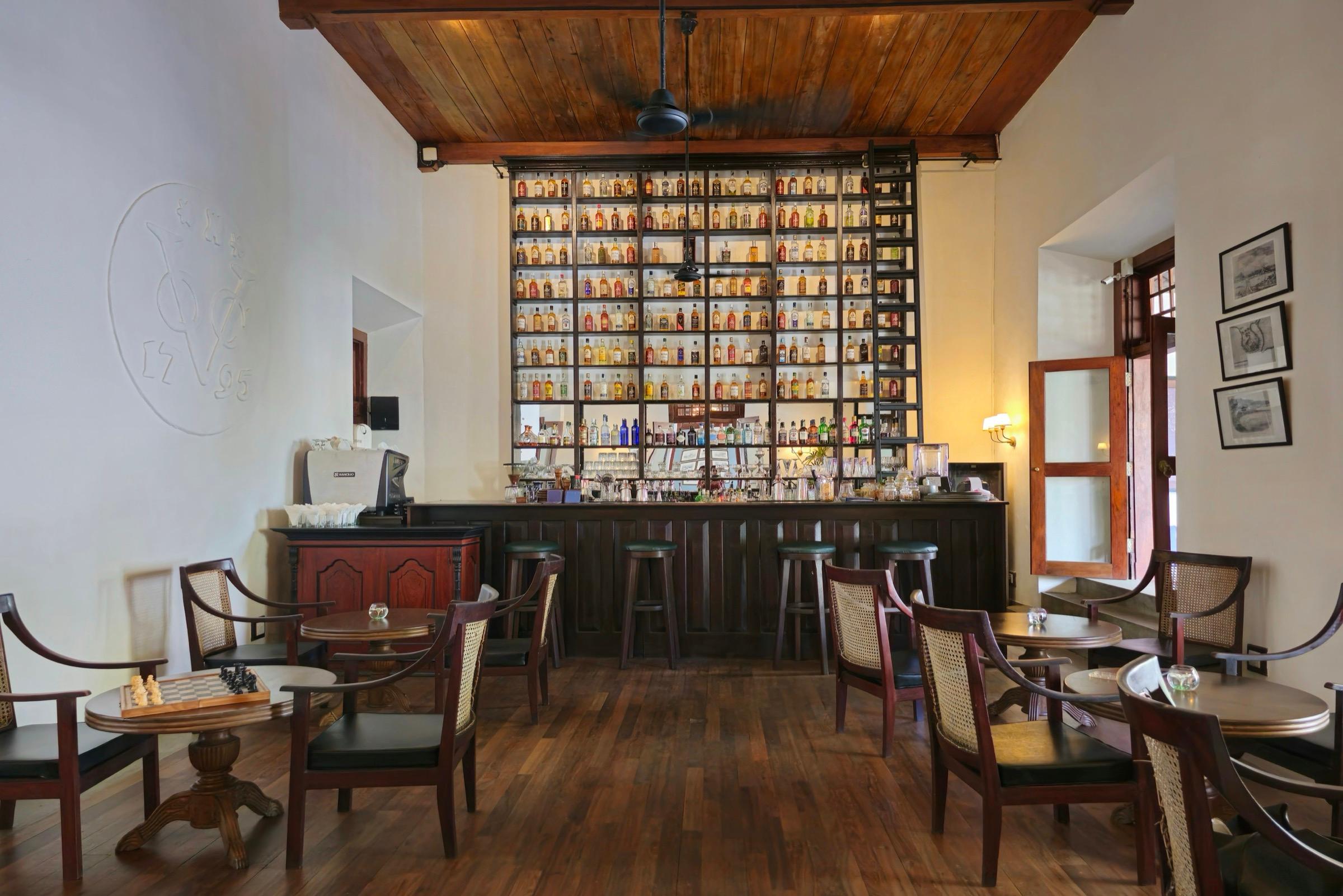 A bar with shelves of spirits lit from below, bar stools along the counter, and cane armchairs around tables set with a chess board, under a wood-beamed ceiling.