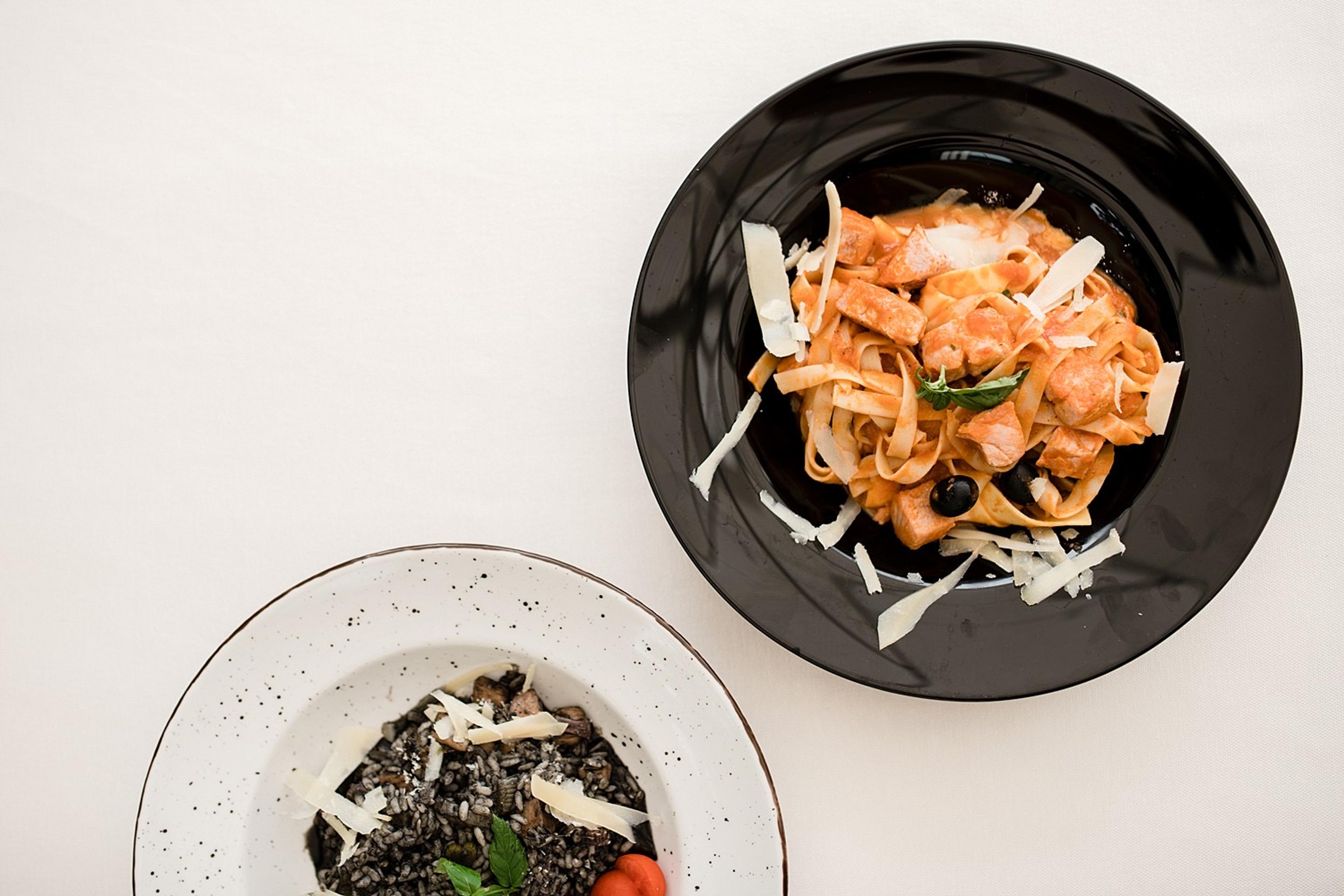 Two plates of food on a white tablecloth, seen from above: a dark plate of pasta with shaved cheese, and a white plate of risotto.