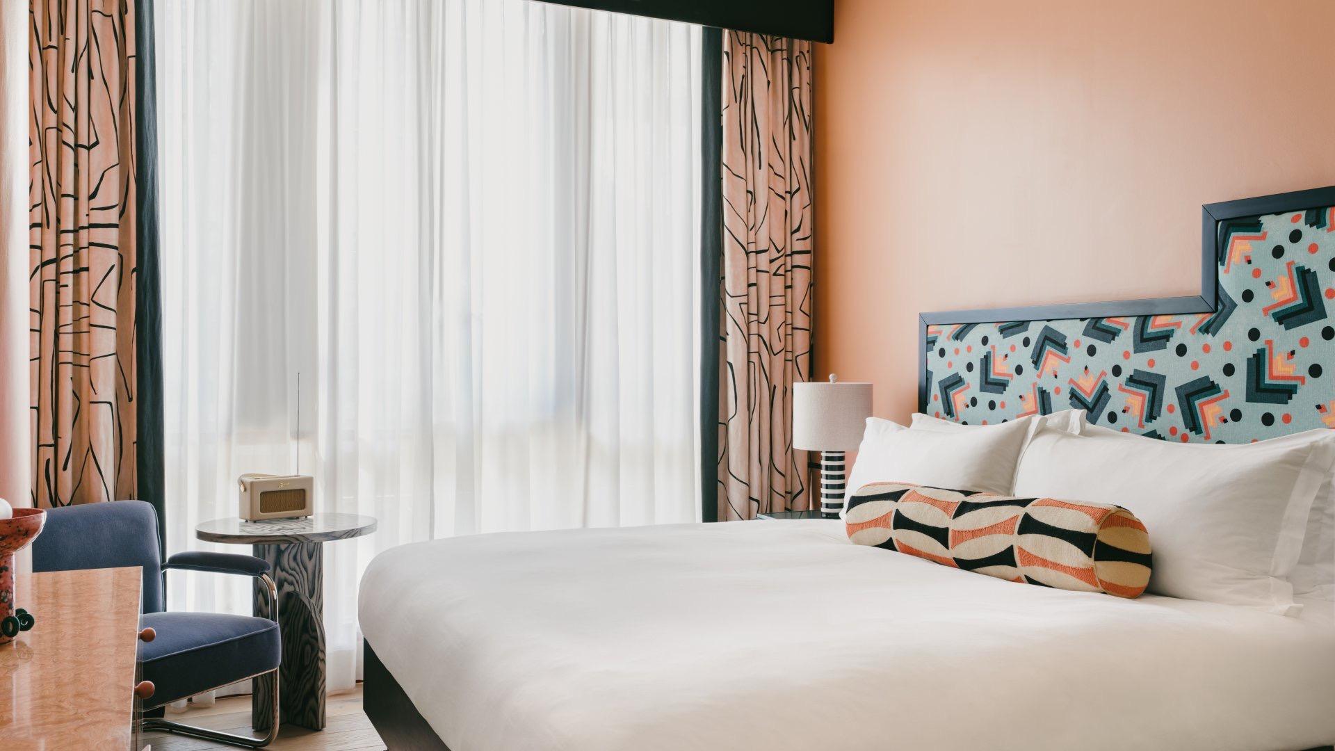 A bedroom with a geometric patterned headboard in teal and orange, peach walls, sheer curtains and a blue armchair at a desk.