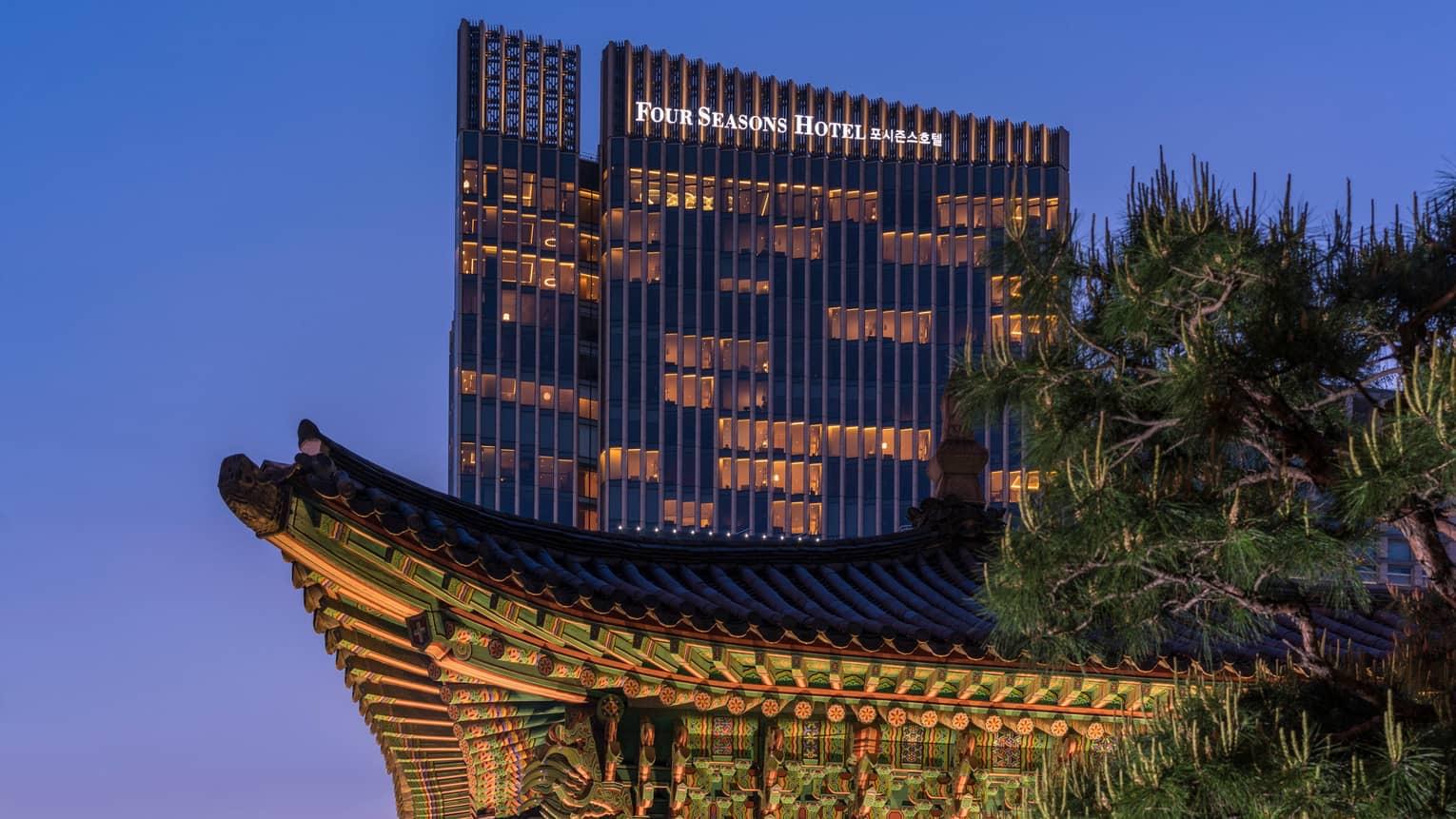 A traditional tiled palace roof in silhouette against a dusk sky, with the lit glass tower of the hotel rising behind it and a pine tree to one side.