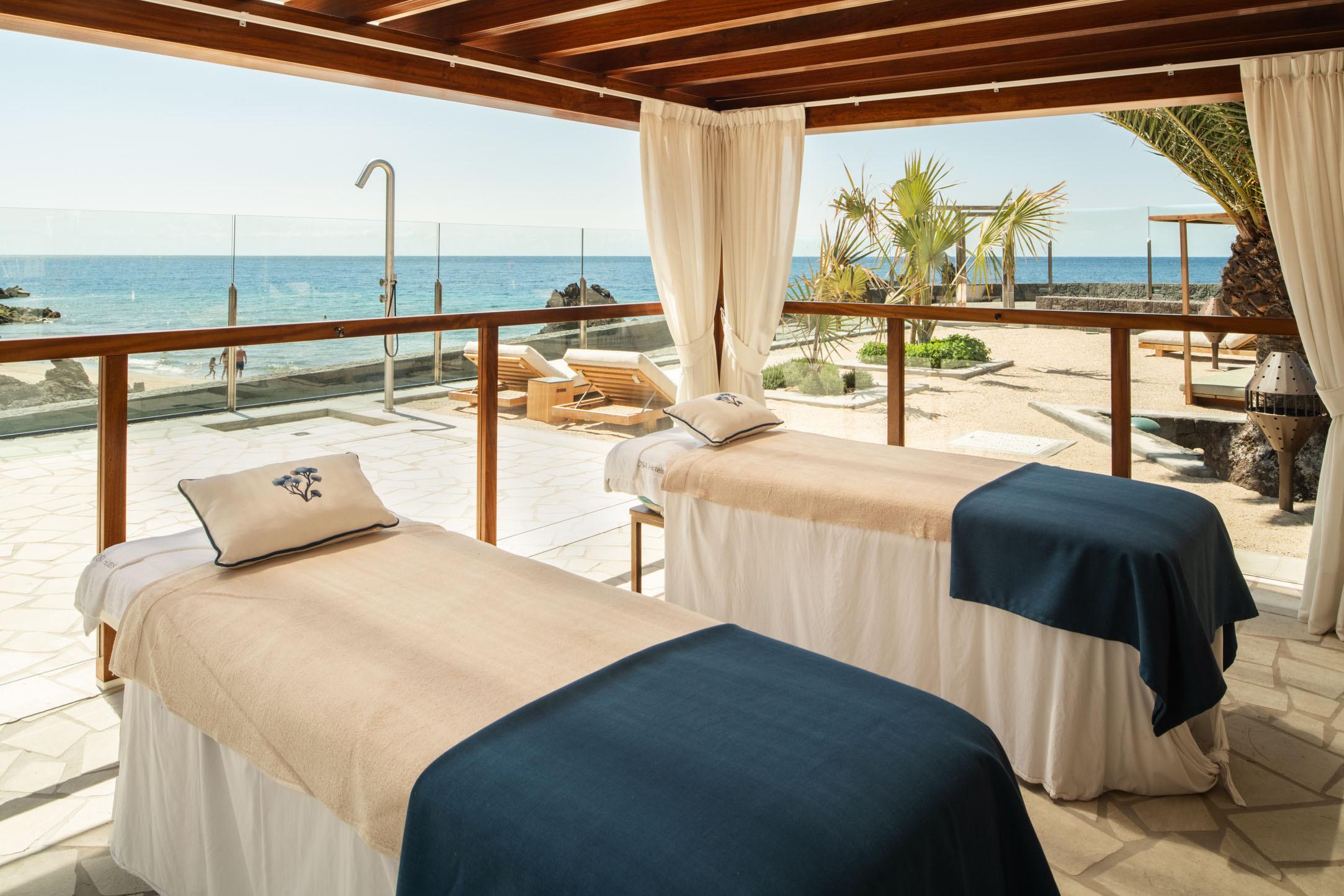 A covered terrace with two treatment beds dressed in cream and navy linen, a railing and curtains framing a view of the sea and palm trees.