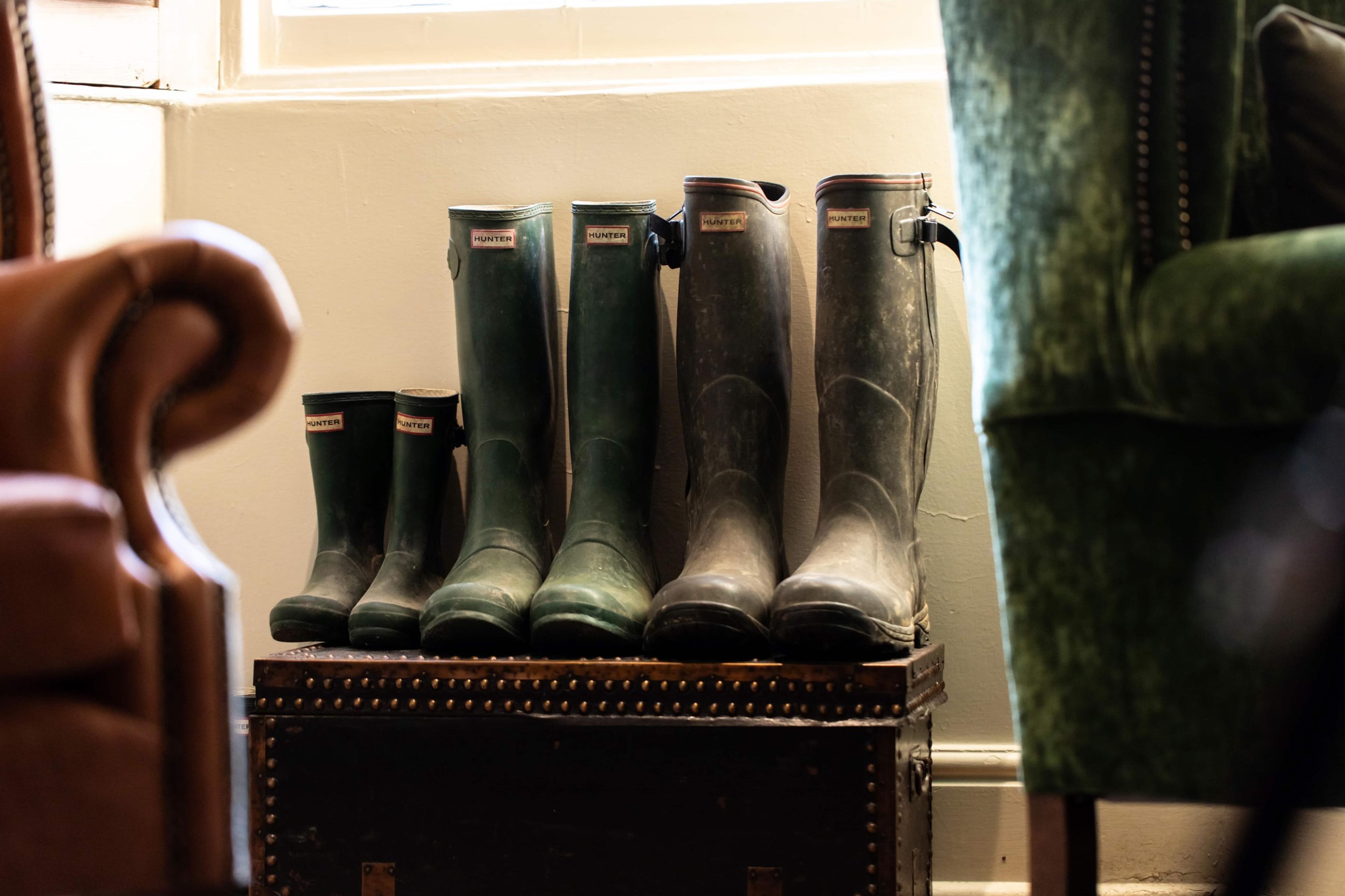 Four pairs of green wellington boots lined up on a trunk by a window, next to a leather armchair.