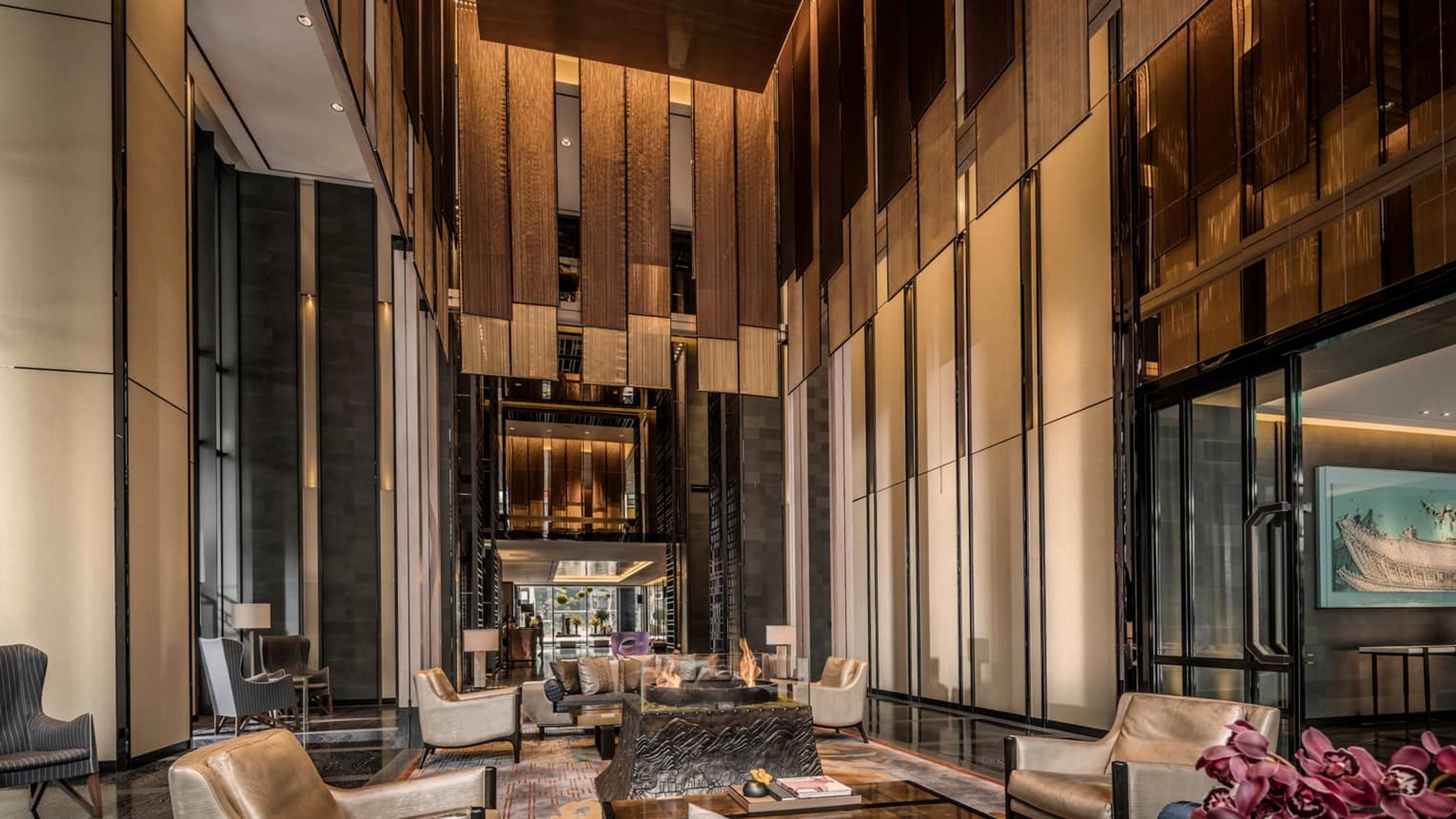 A tall hotel lobby lounge with wood-slat walls, armchairs grouped around a stone table with a lit fire pit, and orchids in the foreground.