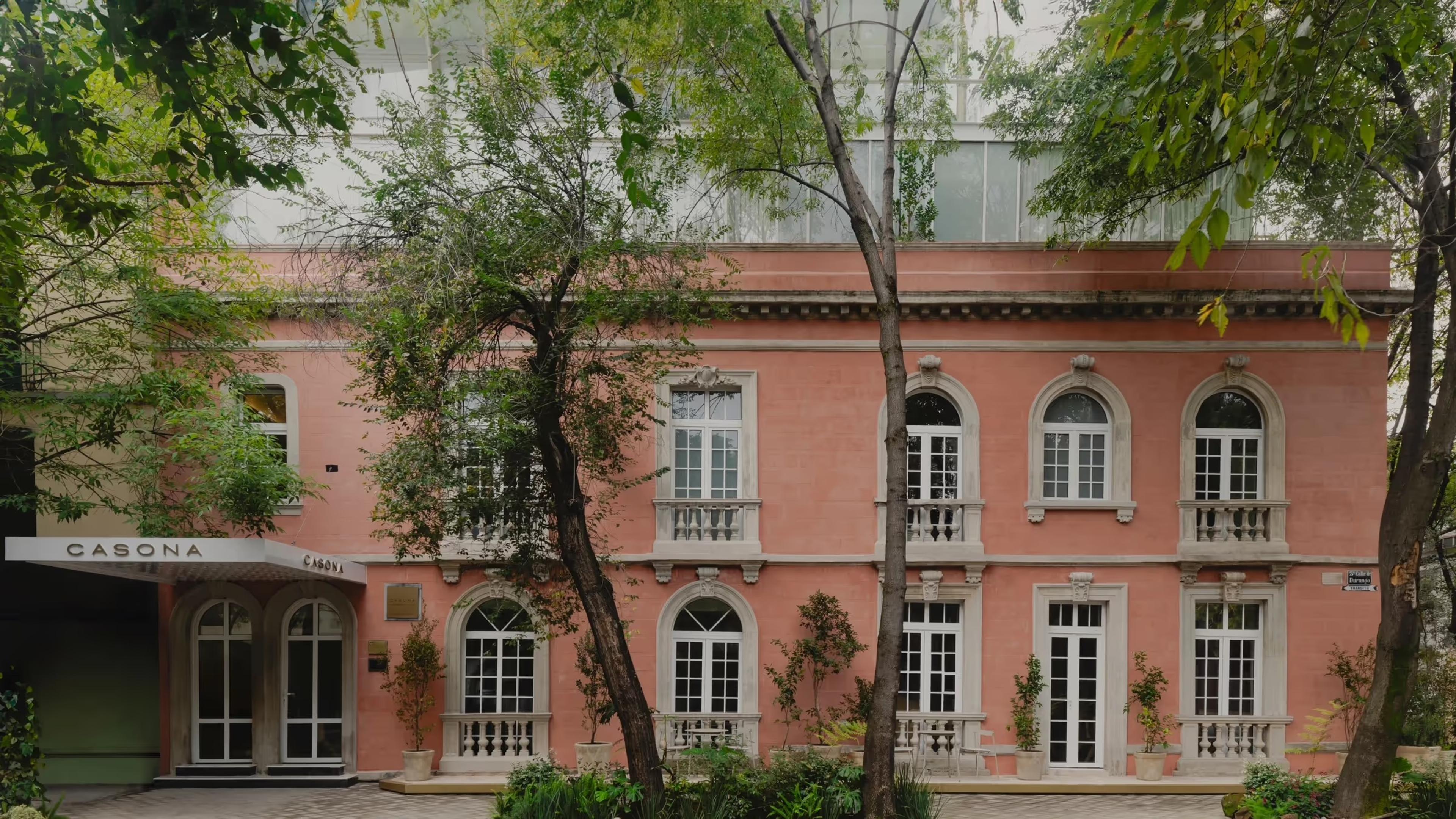 A pink stuccoed colonial mansion with arched windows and iron balconies, its ground-floor glass doors marked Casona, seen through trees.