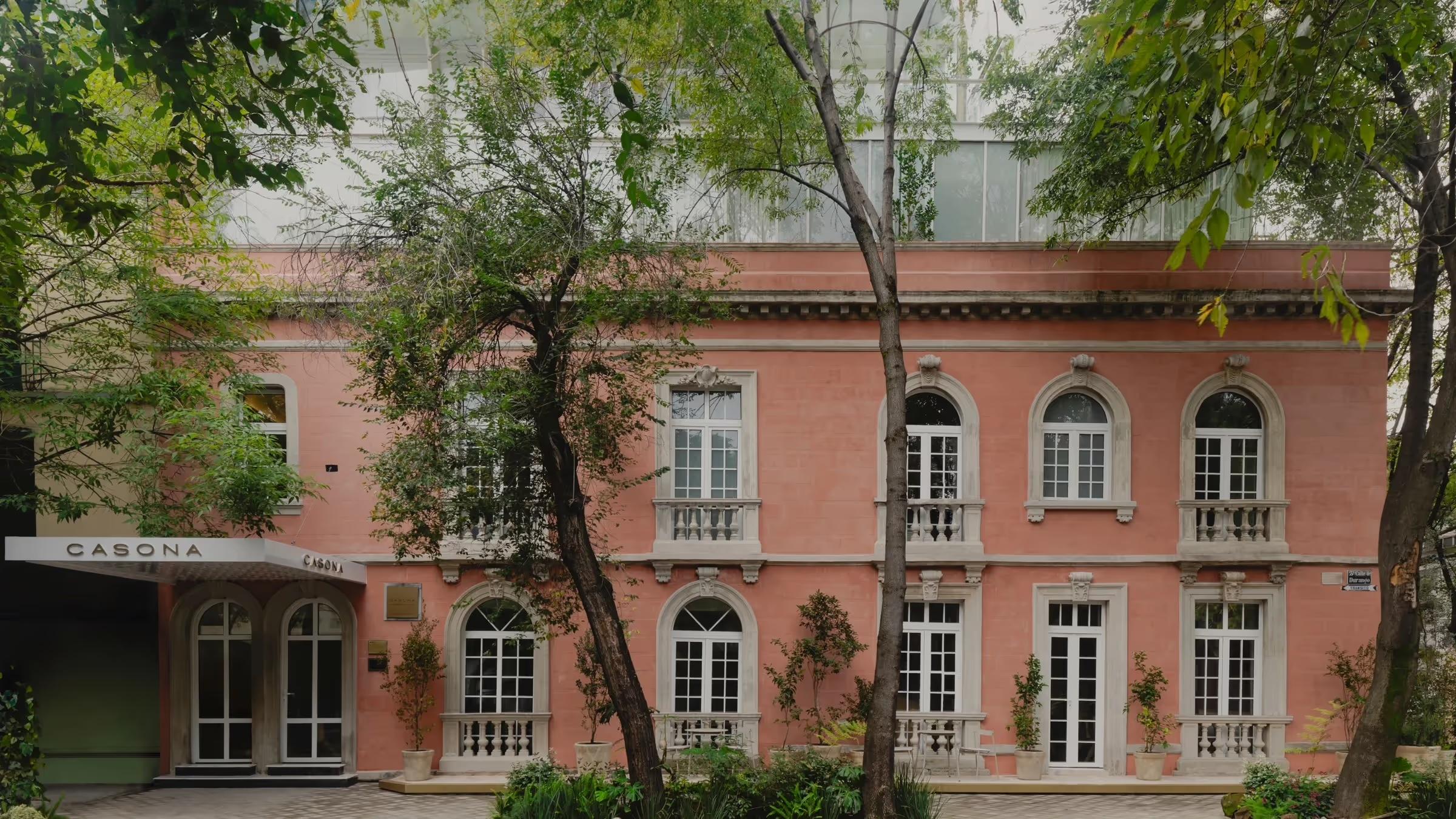 A pink stuccoed colonial mansion with arched windows and iron balconies, its ground-floor glass doors marked Casona, seen through trees.