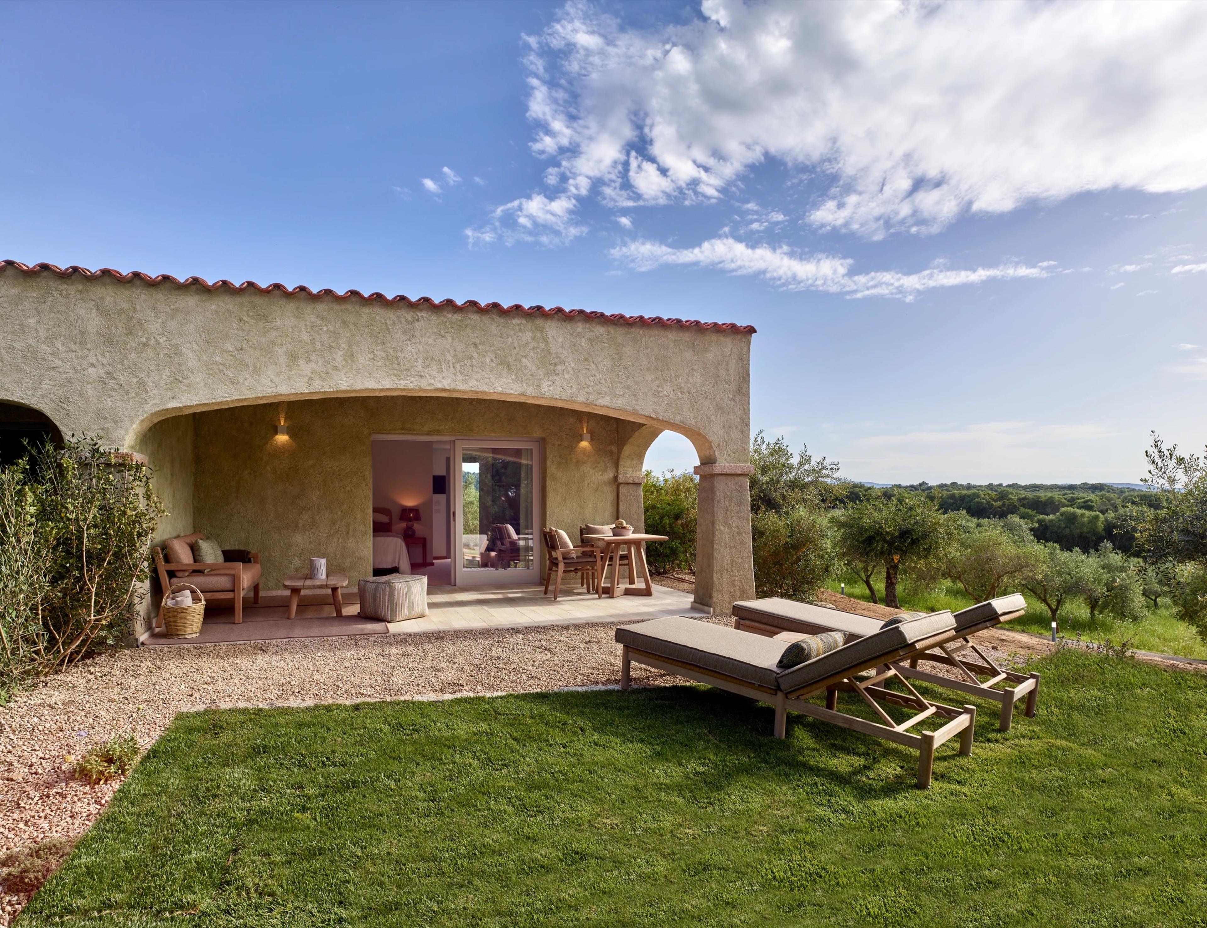 a stone building with a covered terrace furnished with wicker armchairs, two sun loungers on the lawn in front and an olive grove beyond