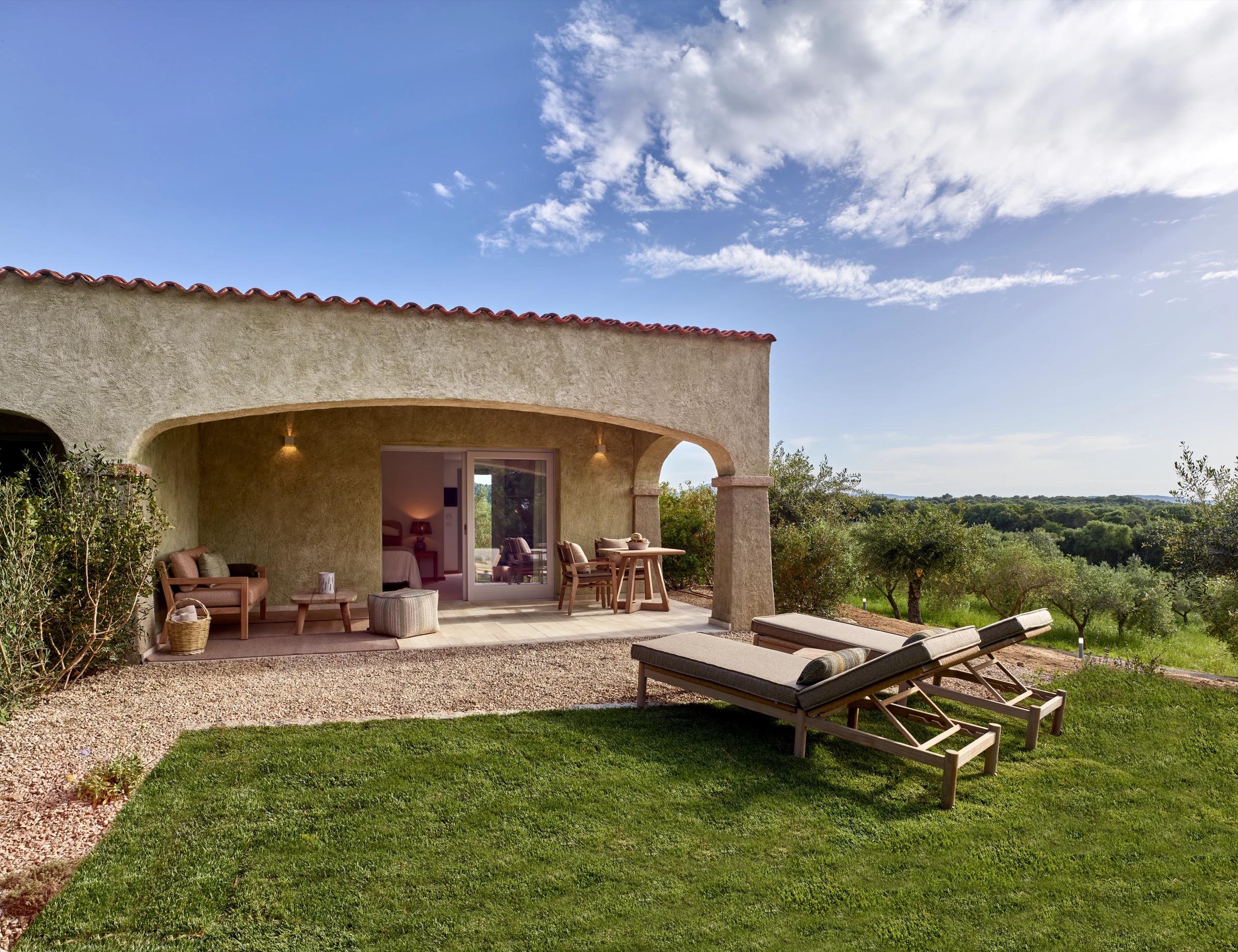 a stone building with a covered terrace furnished with wicker armchairs, two sun loungers on the lawn in front and an olive grove beyond