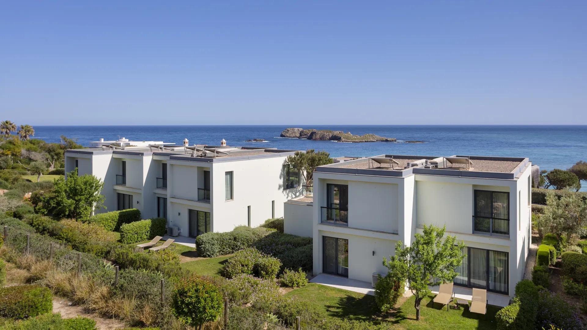 An aerial view of two rows of white flat-roofed houses on a grassy slope, with the sea and a rocky islet behind.