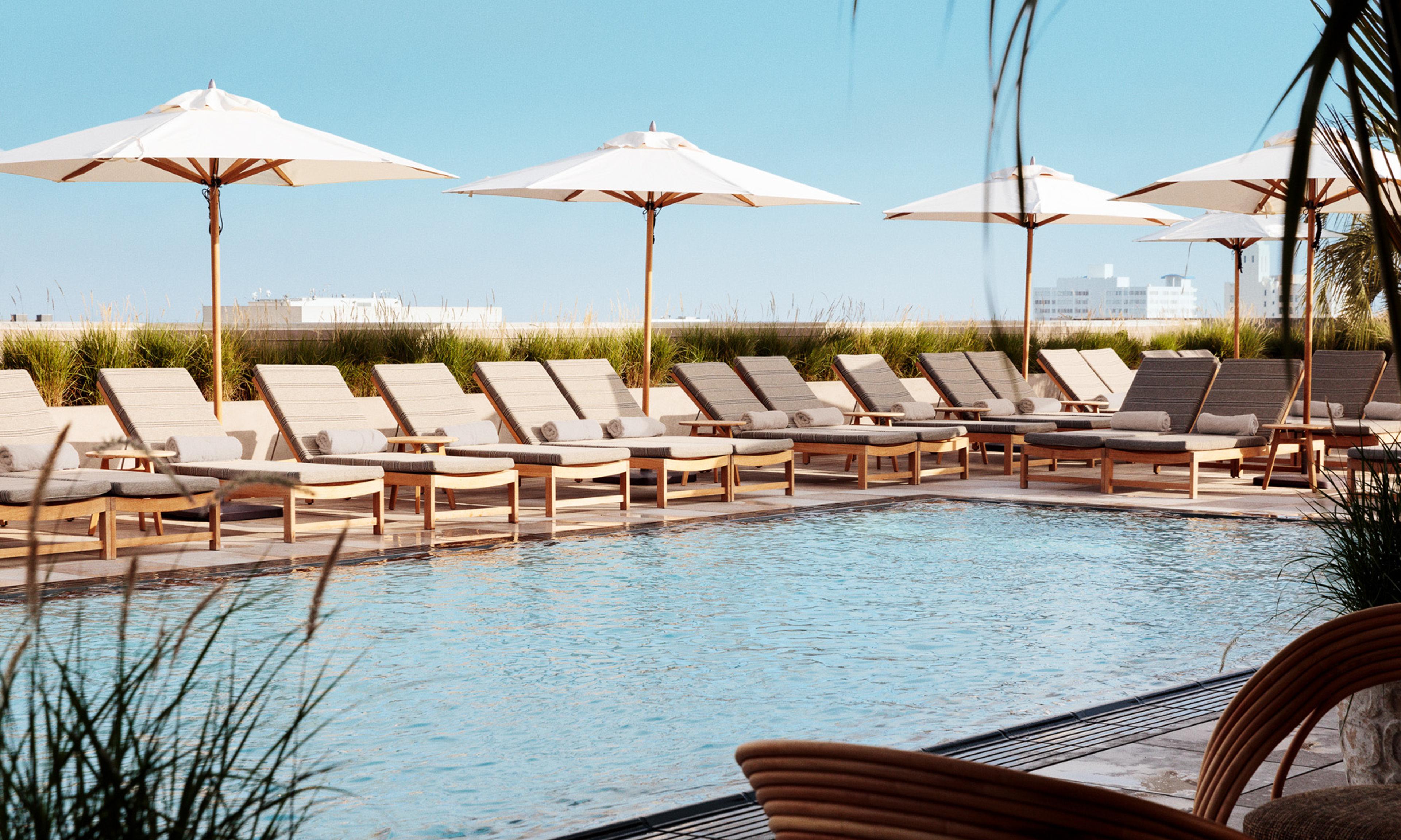 A rooftop pool lined with loungers and white umbrellas, with low grasses at the edge and the city skyline beyond.