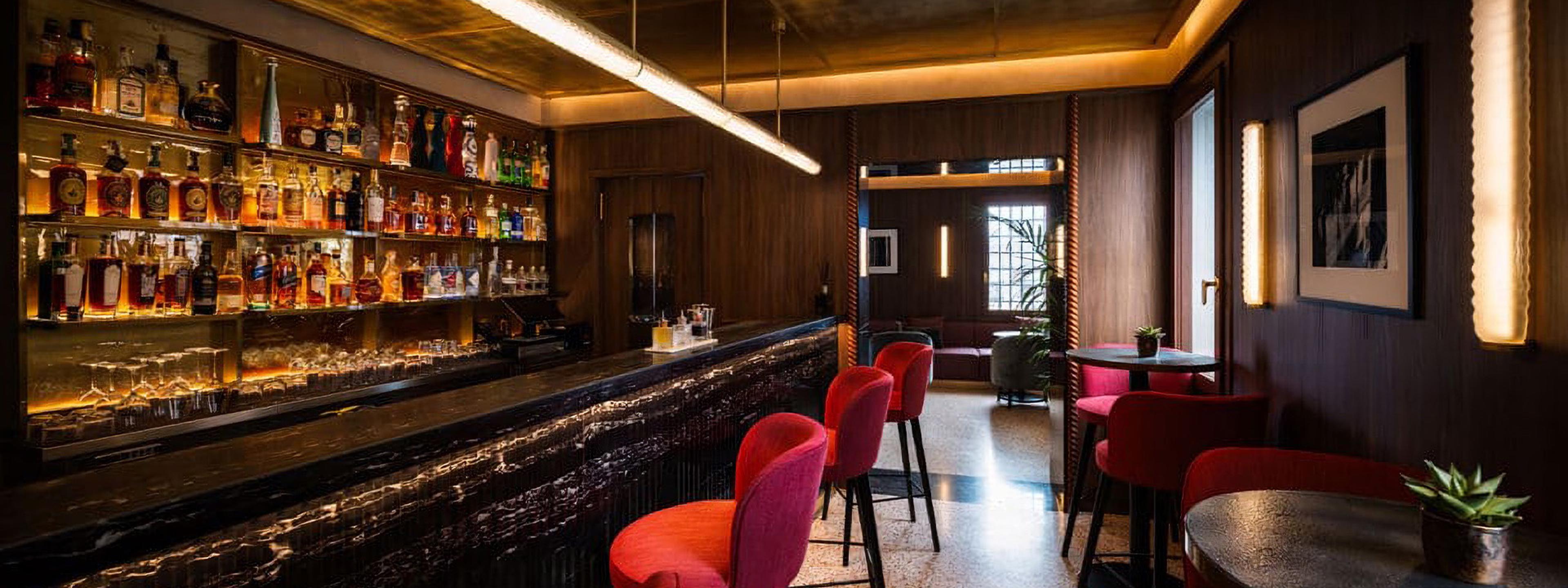 An empty bar room with a marble counter, red upholstered stools, shelves of bottles, and a seating area beyond.
