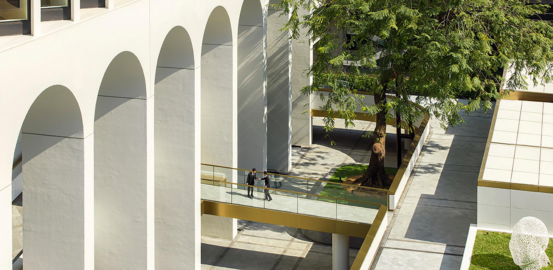 an aerial view of the hotel's arched facade, a glass bridge and a courtyard tree, with two people crossing the bridge