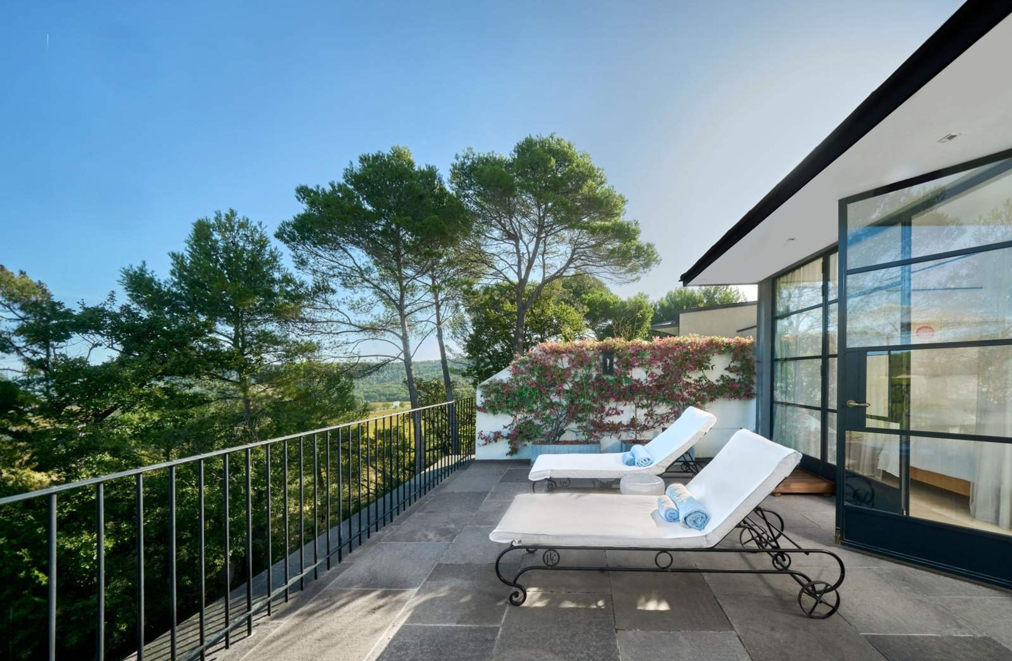 Two chaise loungers with rolled towels on a stone terrace, looking over a wooded valley beside a wall covered in flowering vine.