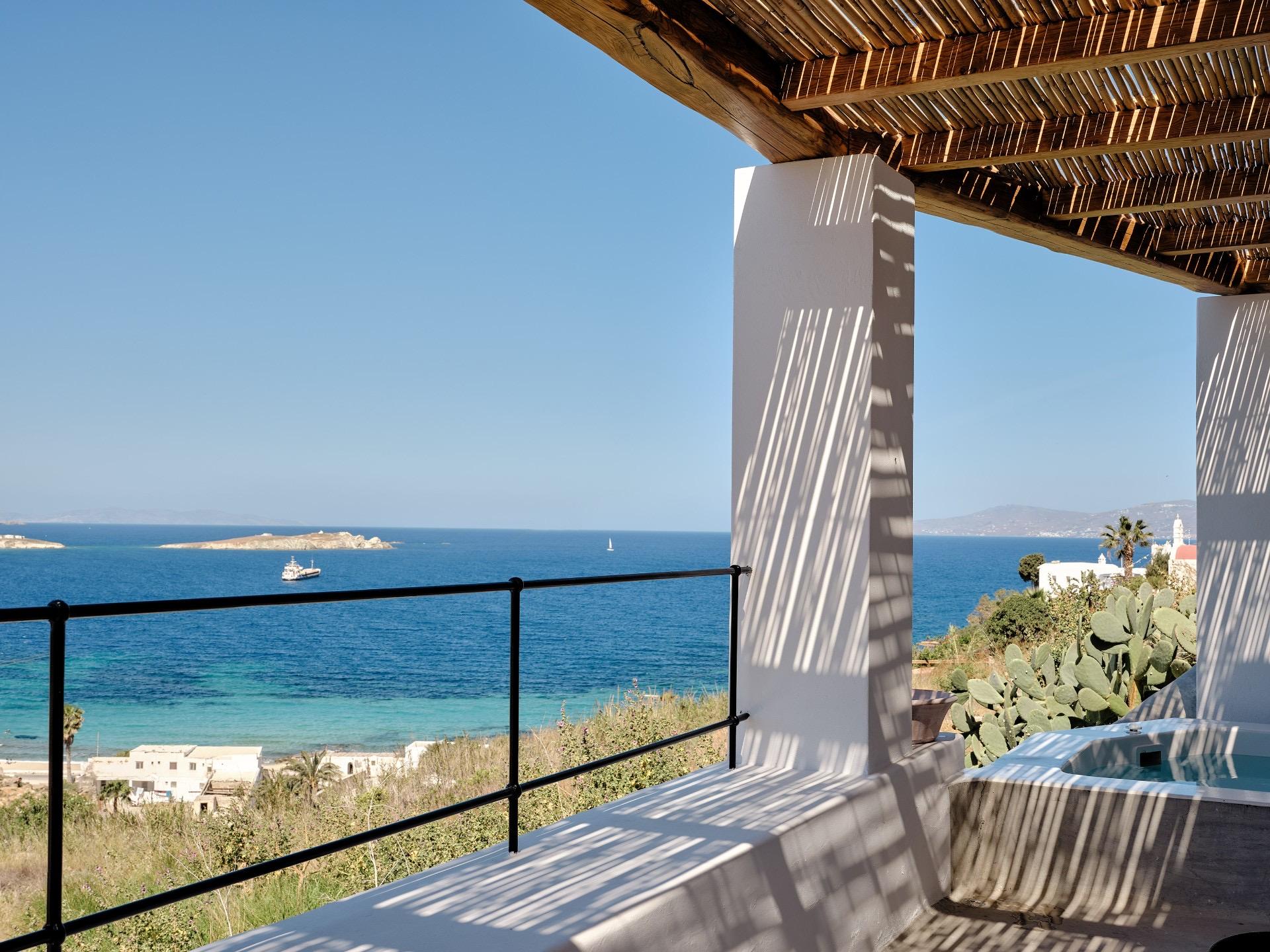 A shaded pergola corner looks out past a plunge pool to the sea, a boat on the water and a hillside village in the distance.