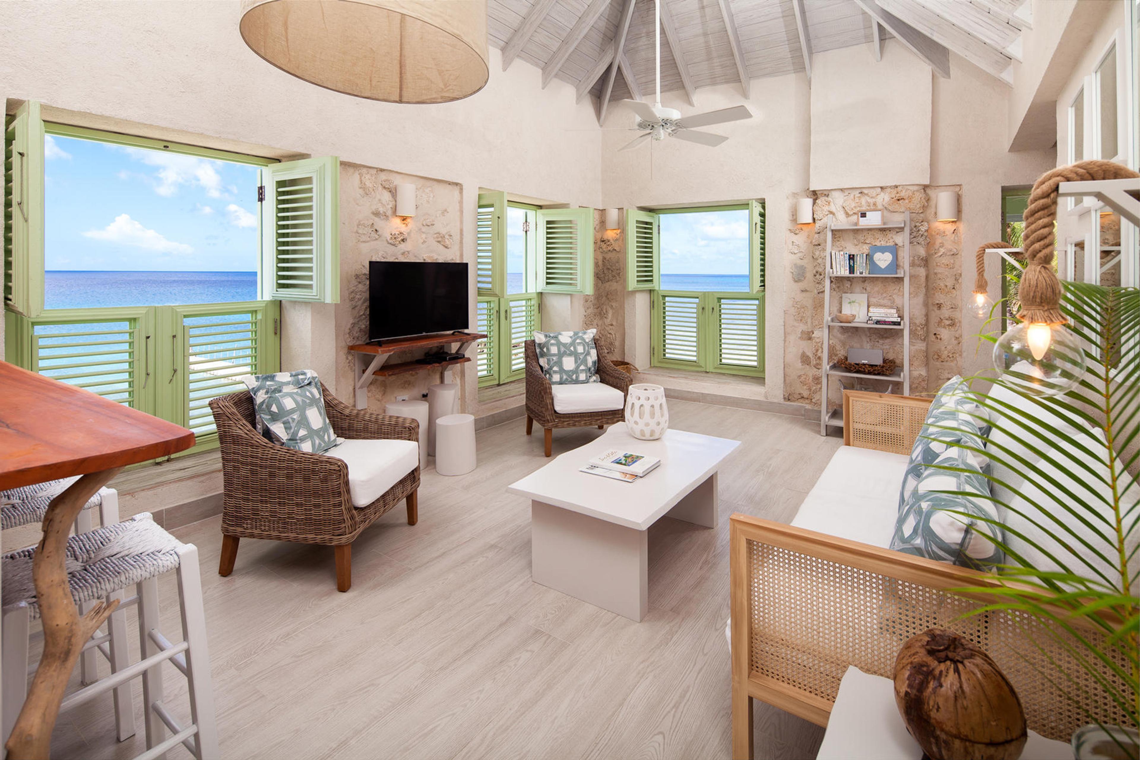 A living room with wicker armchairs and a sofa, green shuttered windows open to a wide view of the sea.