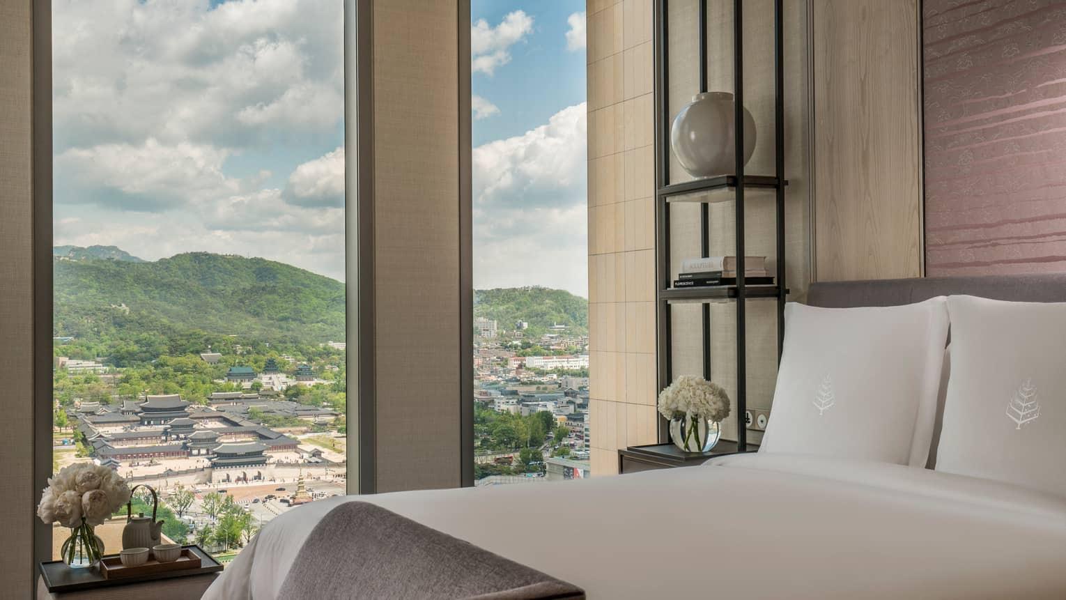 A hotel bed with a padded bench at its foot and a shelving unit beside it, facing floor-to-ceiling windows that look out over a palace complex and green hills.