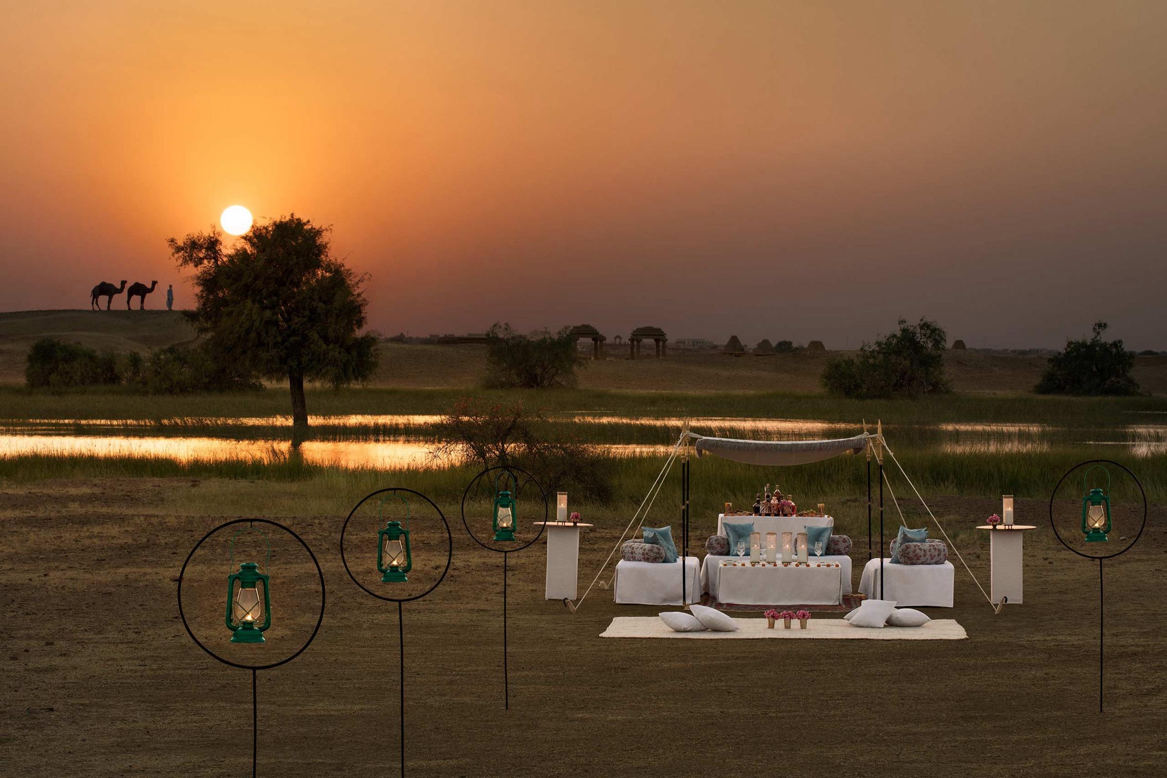 a low table laid for dinner in the desert at sunset, with two camels and their handler silhouetted on a dune behind